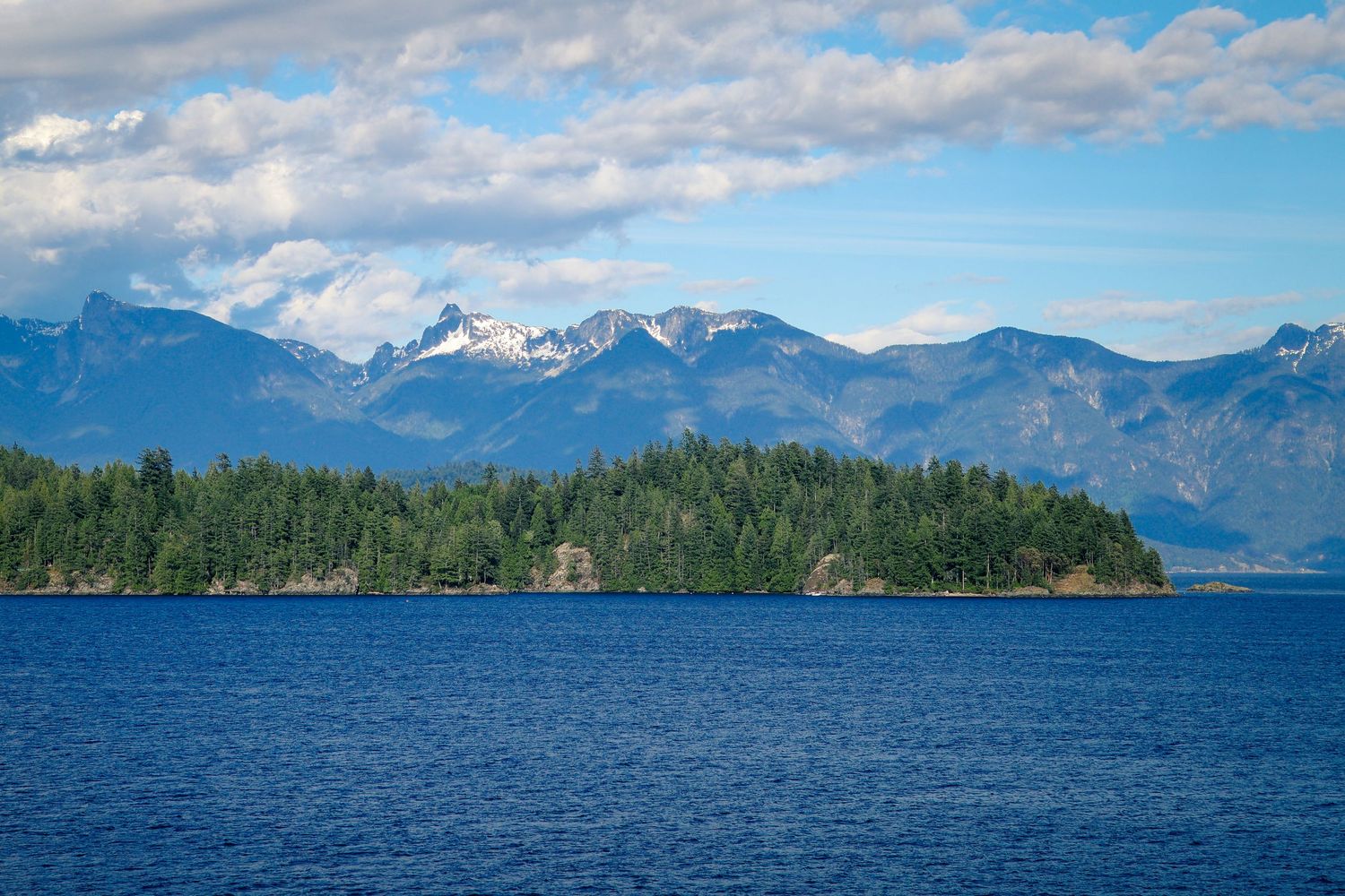 Scenic view of deep blue ocean waters with forested islands and snow-capped mountain peaks under a partly cloudy sky.
