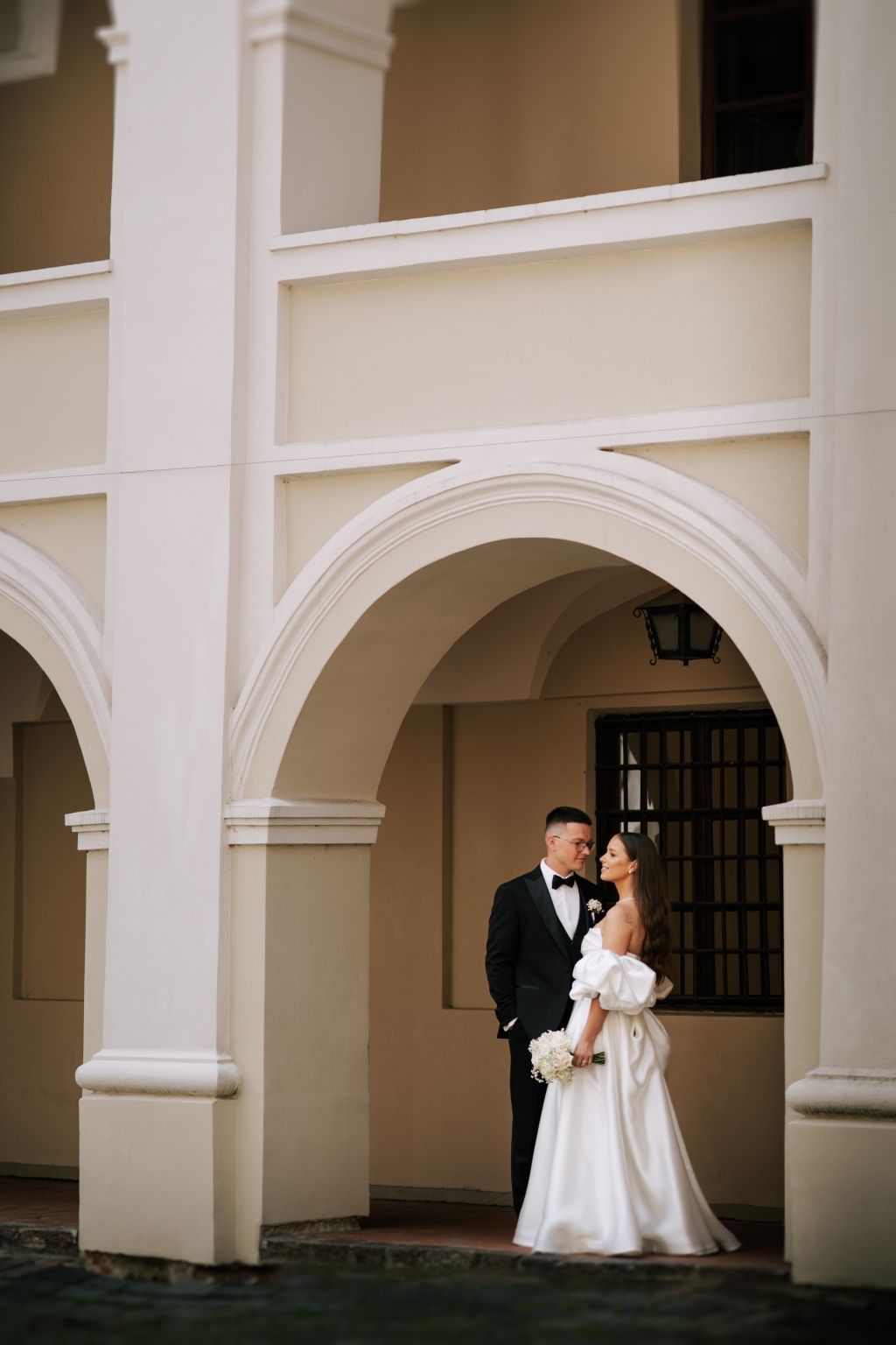 A wedding couple shares an intimate moment under white arched columns in a classical building.