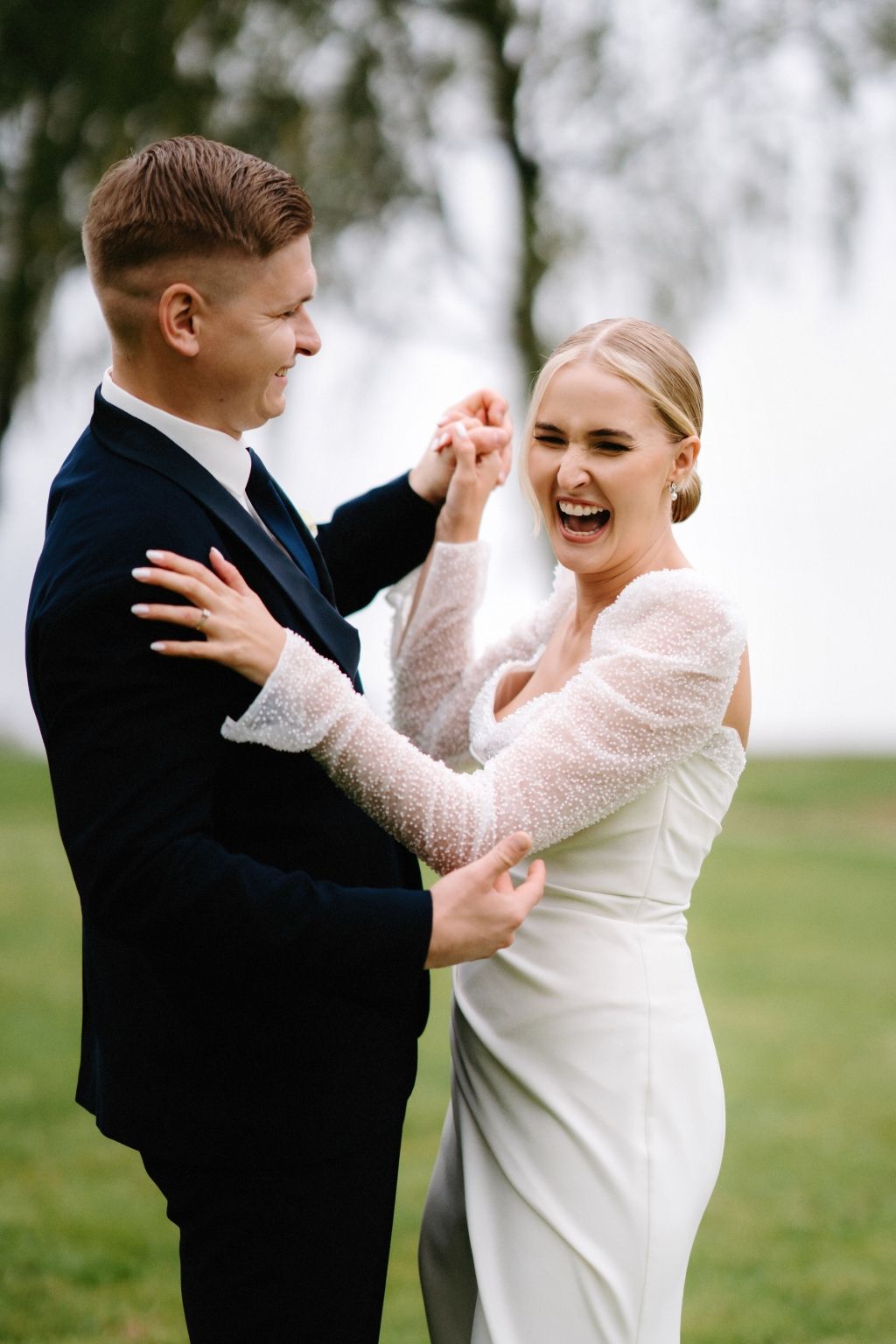 Joyful couple in wedding attire dancing and laughing together on grassy field.