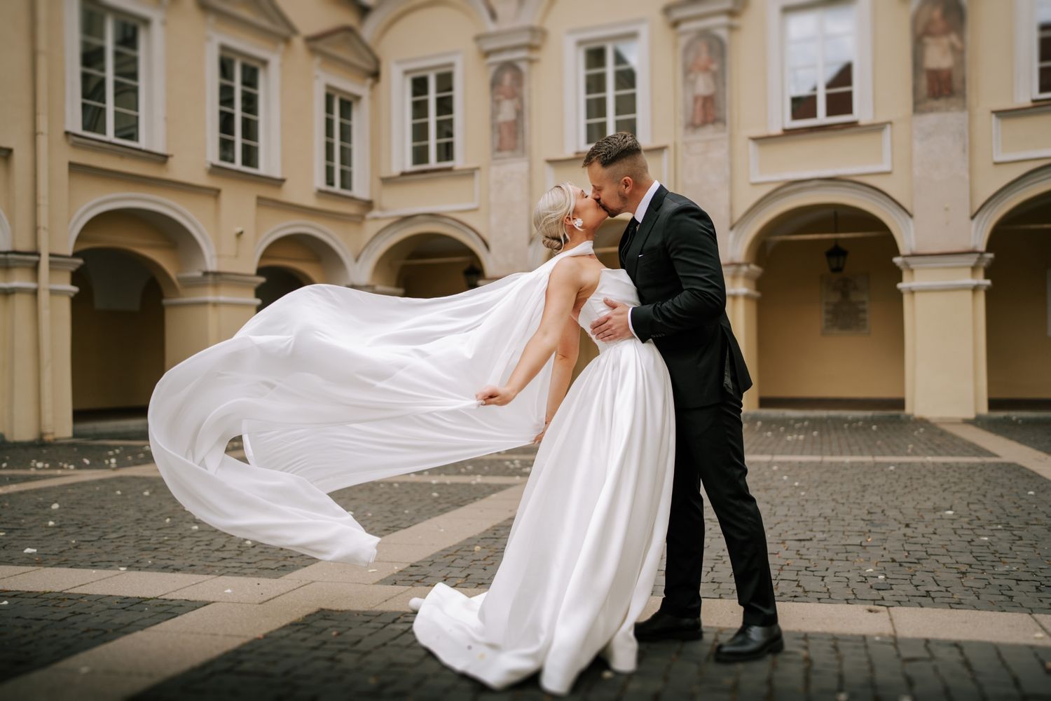 Wedding couple playing with flowing dress in historic courtyard setting.