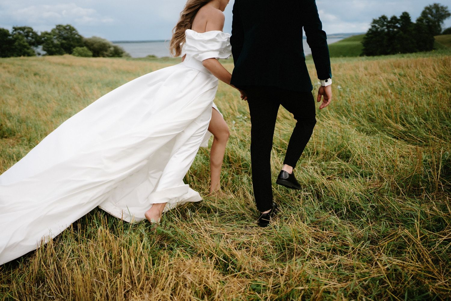 A wedding dress flows dramatically in wind as couple walks through tall grass.