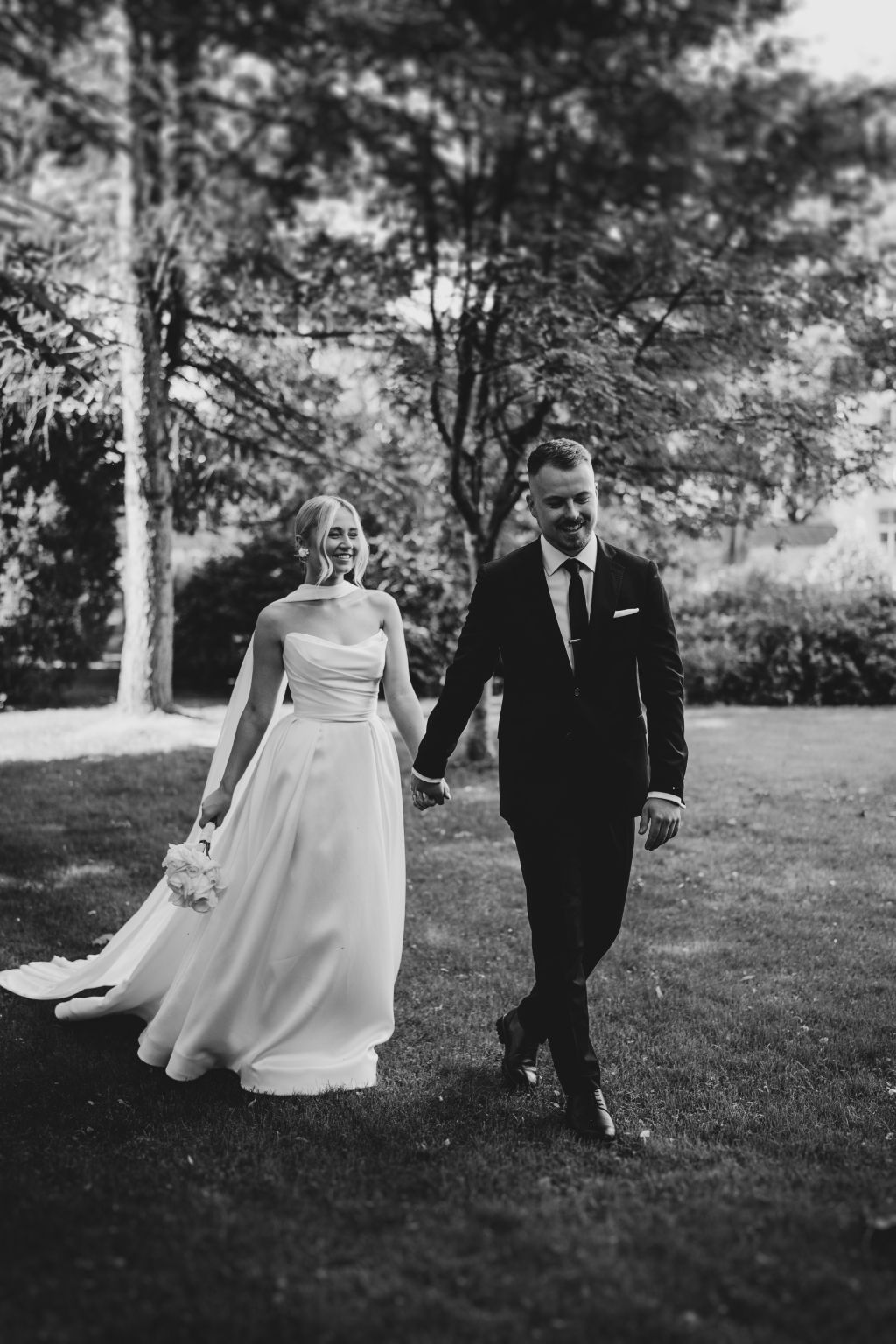 Black and white photo of newlyweds walking together on a grassy lawn.