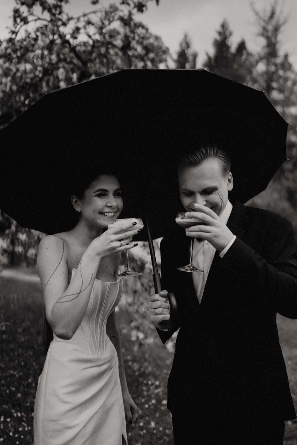 A couple in wedding attire shares drinks under large black umbrellas in a garden setting.