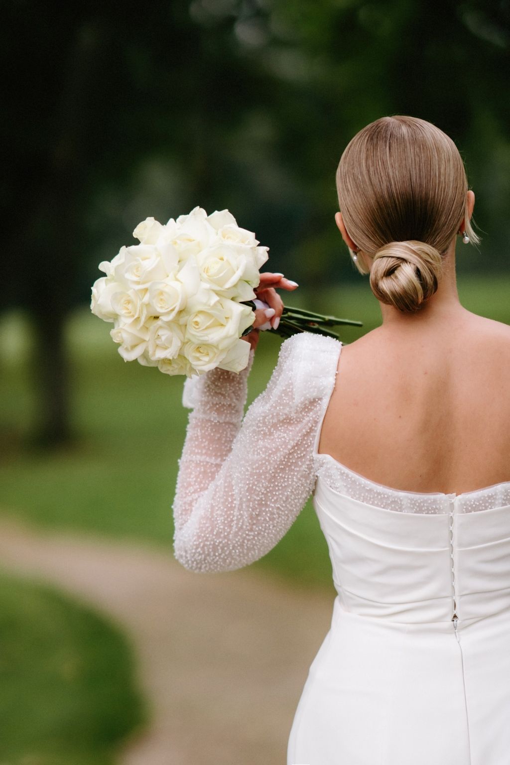 Bride in long-sleeved white dress holding bouquet of white roses from behind.