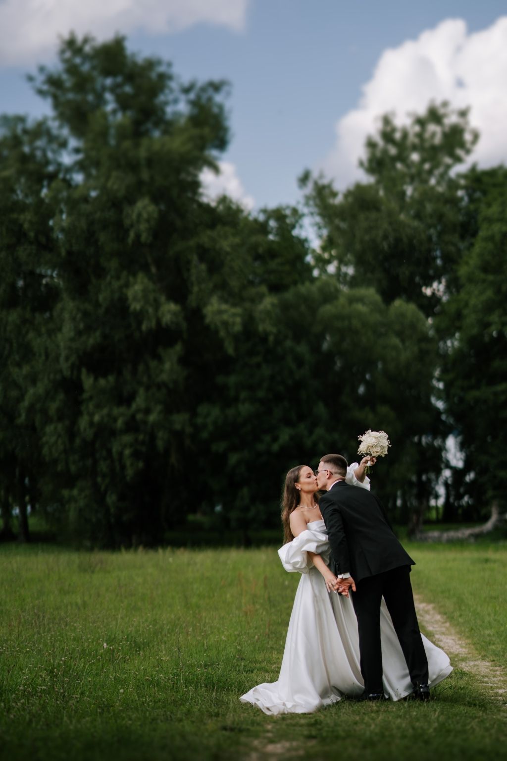 A wedding couple shares romantic moments in a lush green field on a cloudy summer day.