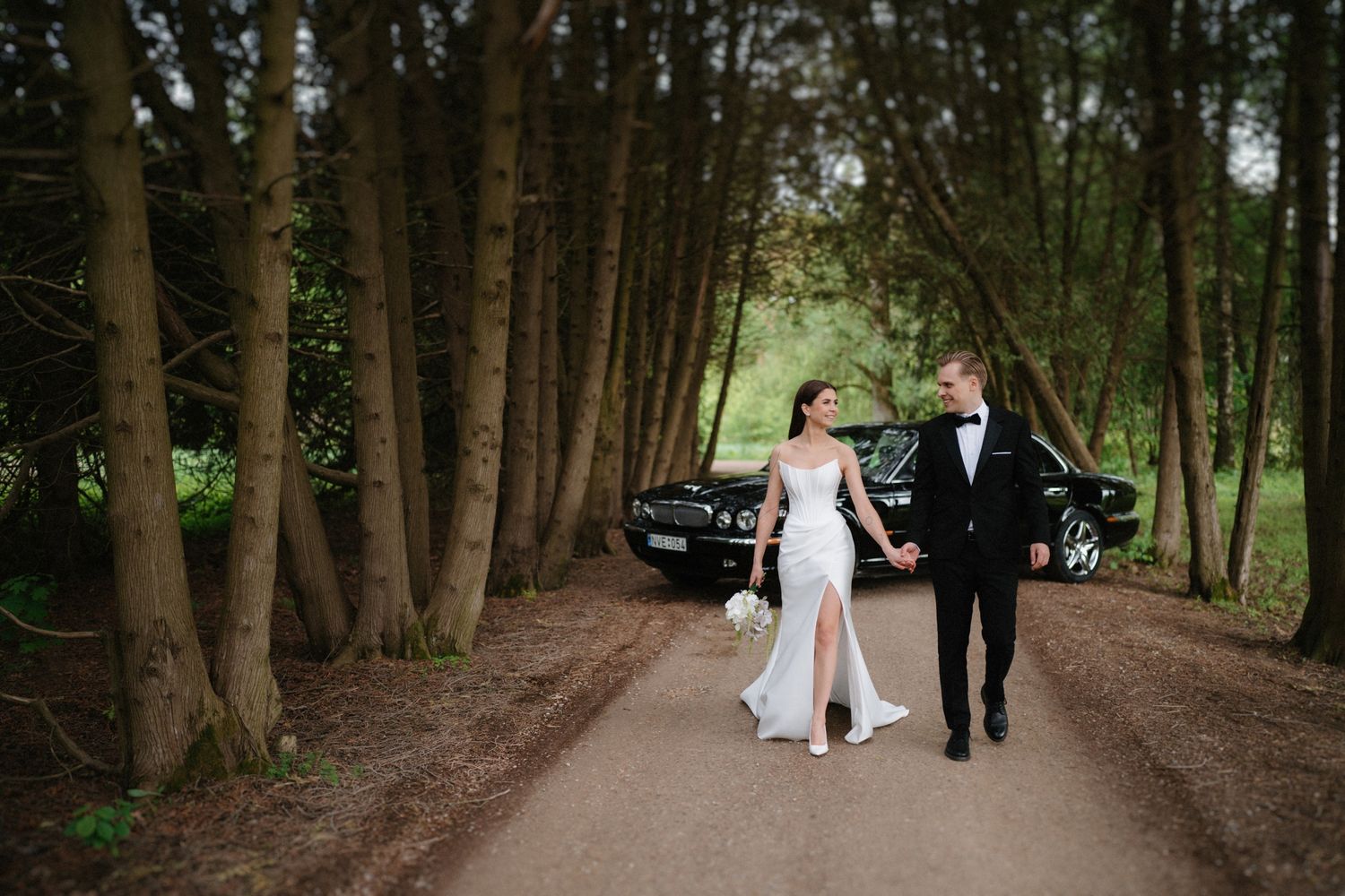 A couple walks hand in hand on a tree-lined path with a classic car in the background.
