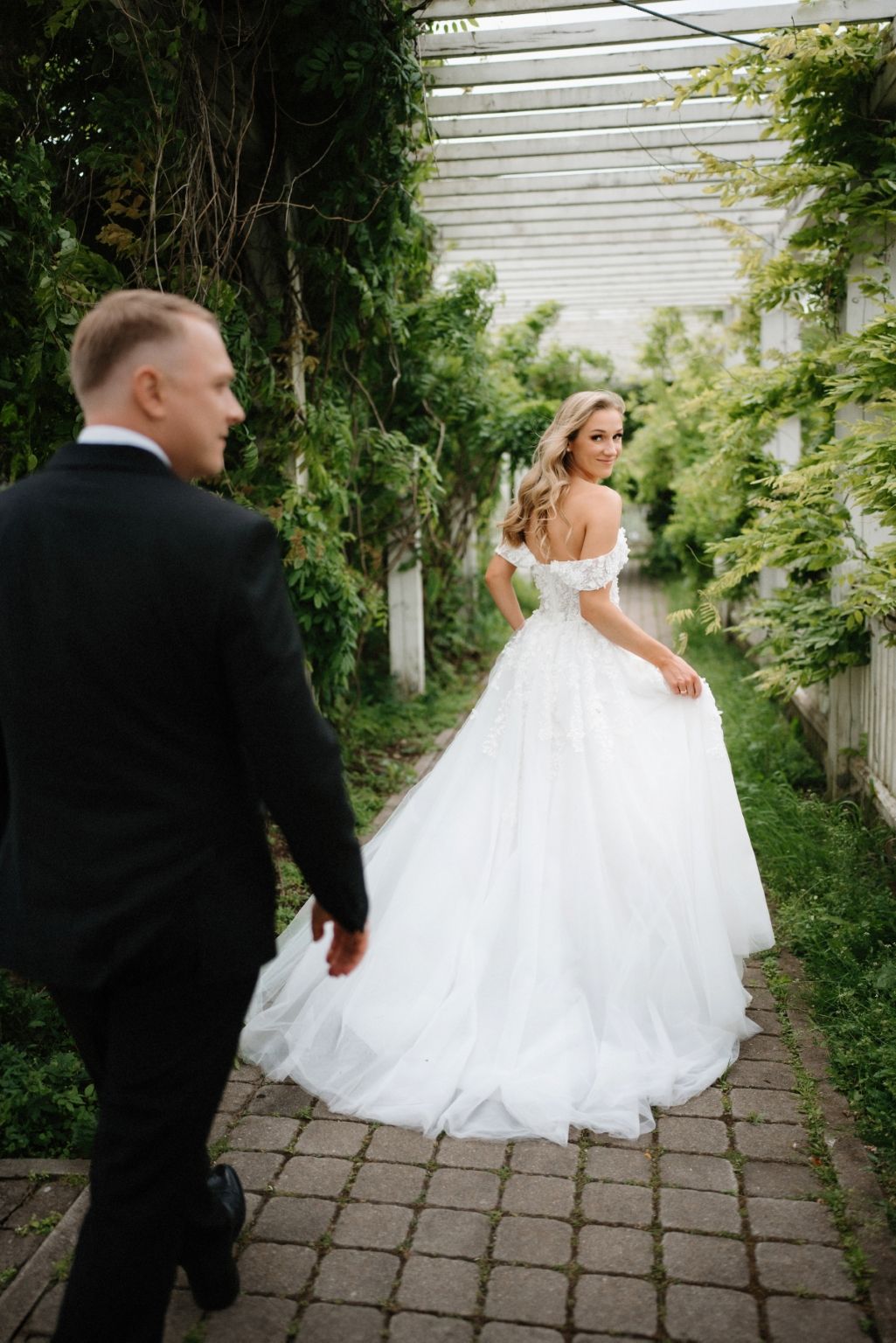 Wedding couple walking along a garden path with white fence and greenery.