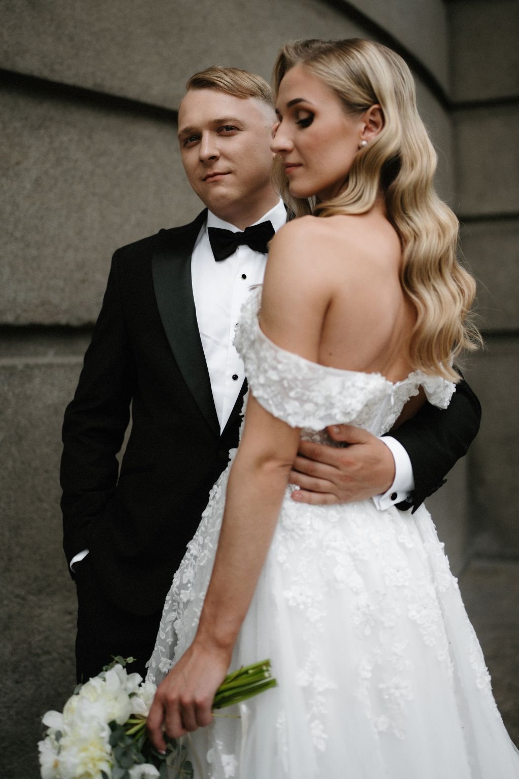 Bride in off-shoulder lace gown and groom in tuxedo embrace during outdoor wedding photos.