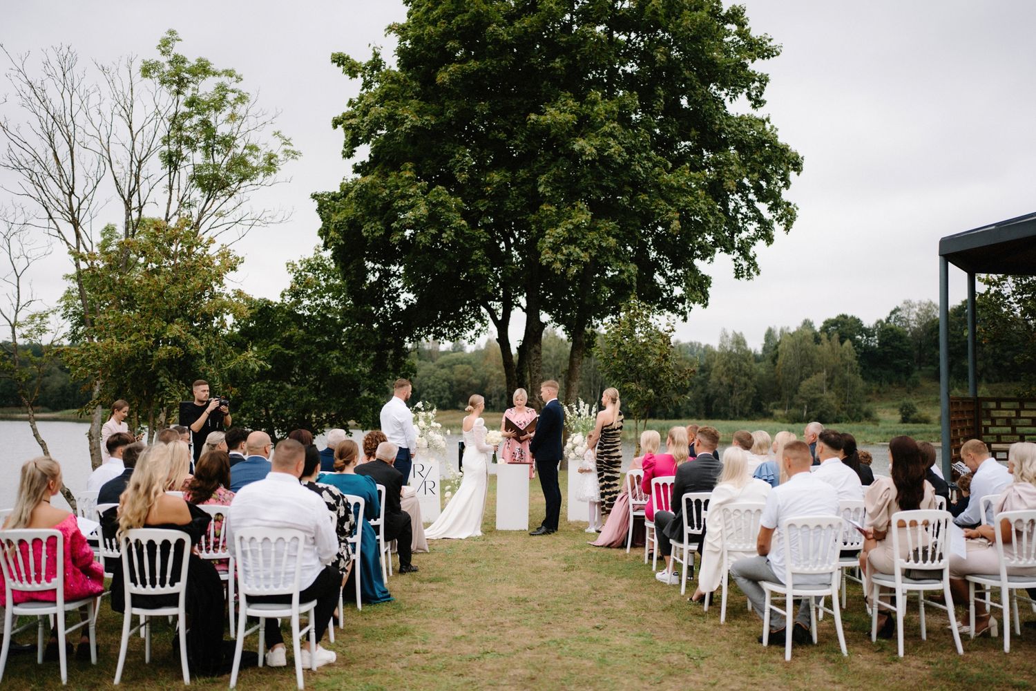 Guests seated in white chairs attend an outdoor lakeside wedding ceremony.