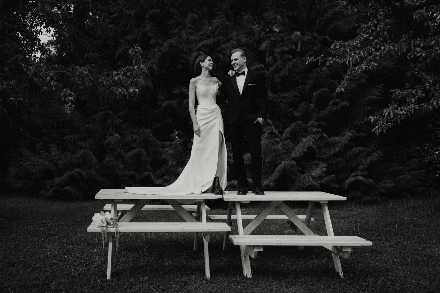 A couple in formal wear stands playfully on a picnic table outdoors.