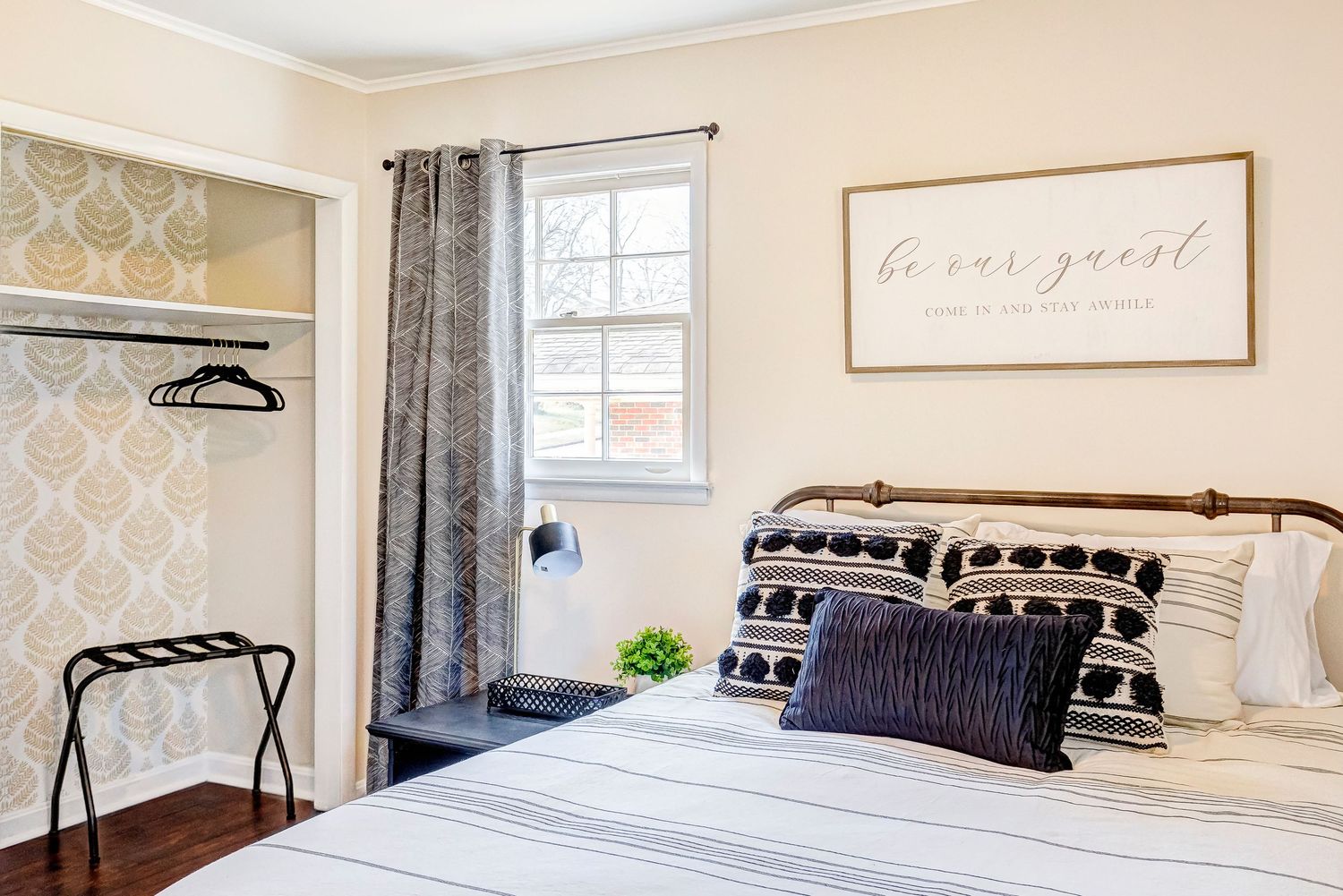 A cozy guest bedroom with neutral decor features a bed with striped pillows, window with gray curtains, and live love laugh wall art.