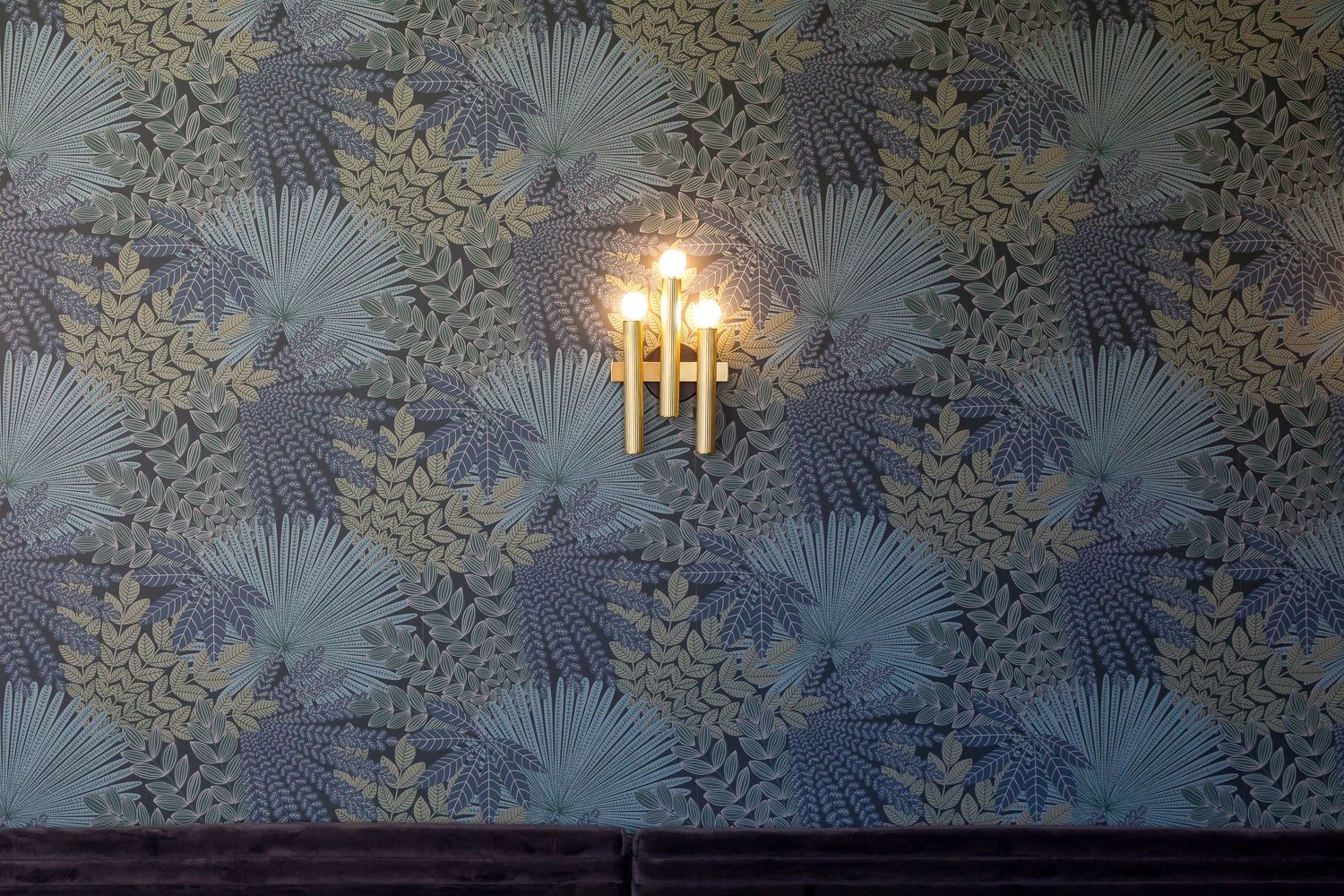 A vintage brass wall sconce illuminates a blue floral wallpaper with palm frond and botanical pattern.