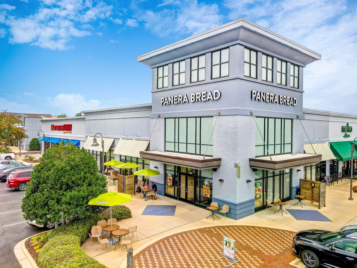Modern retail shopping center with Paper Source store and outdoor seating beneath yellow umbrellas on curved walkway.