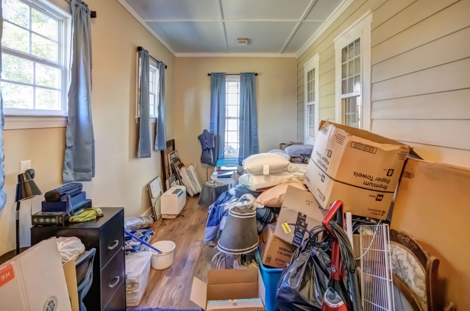 A cluttered room with moving boxes, household items, and hardwood floors near a window with blue curtains.