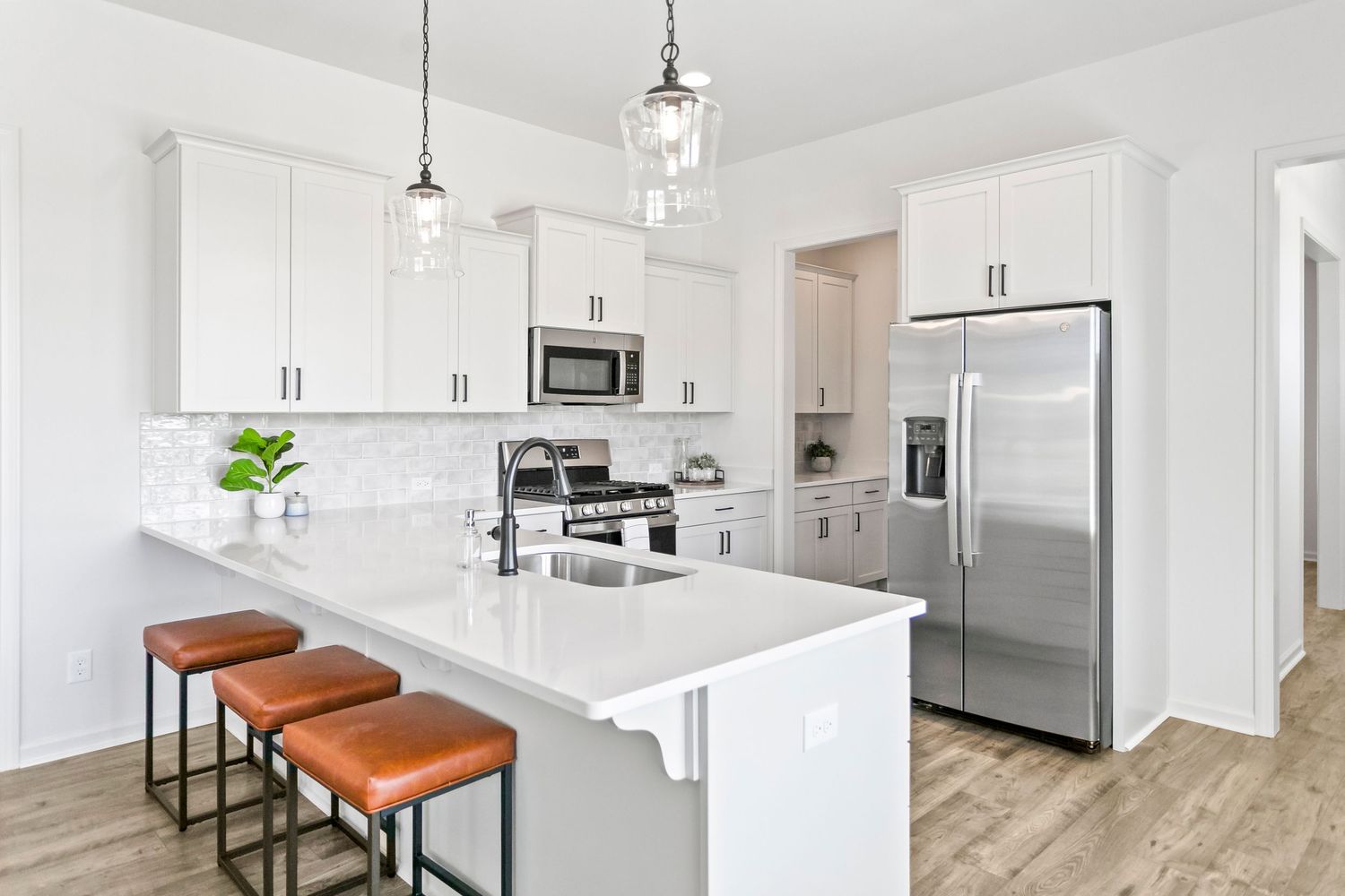 Modern white kitchen with quartz countertops, brown leather barstools, and stainless steel appliances.