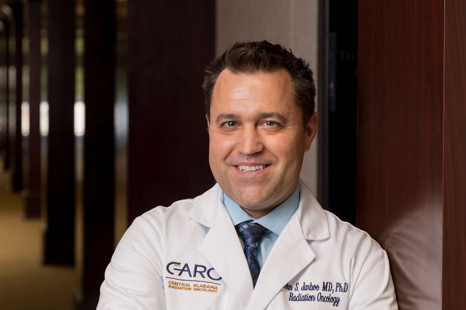 Medical professional in white coat and blue tie stands in professional office setting with dark wood paneling.
