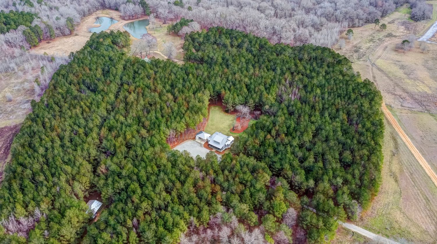 Aerial drone view of a house with swimming pool surrounded by dense green forest in a rural setting.