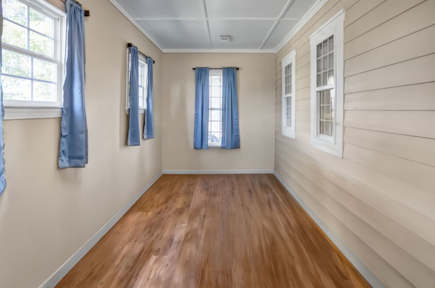 Empty room interior, clean with beige walls, hardwood flooring, and multiple windows providing natural light.