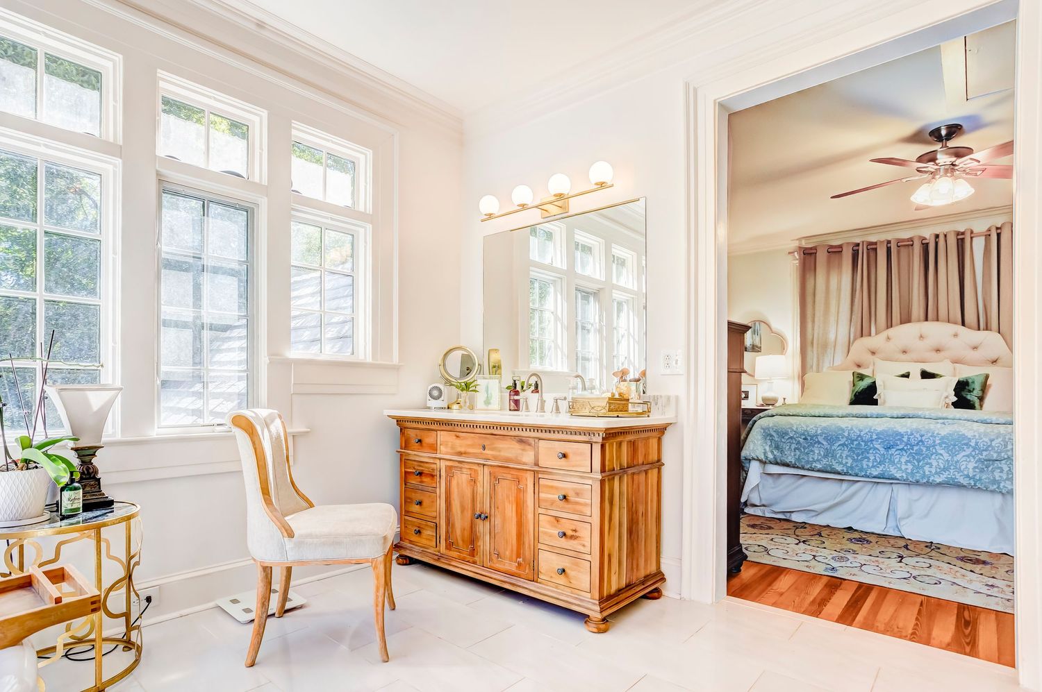 An elegant bathroom with a wooden vanity, gold mirror, white walls, and a view into a bedroom with blue bedding.