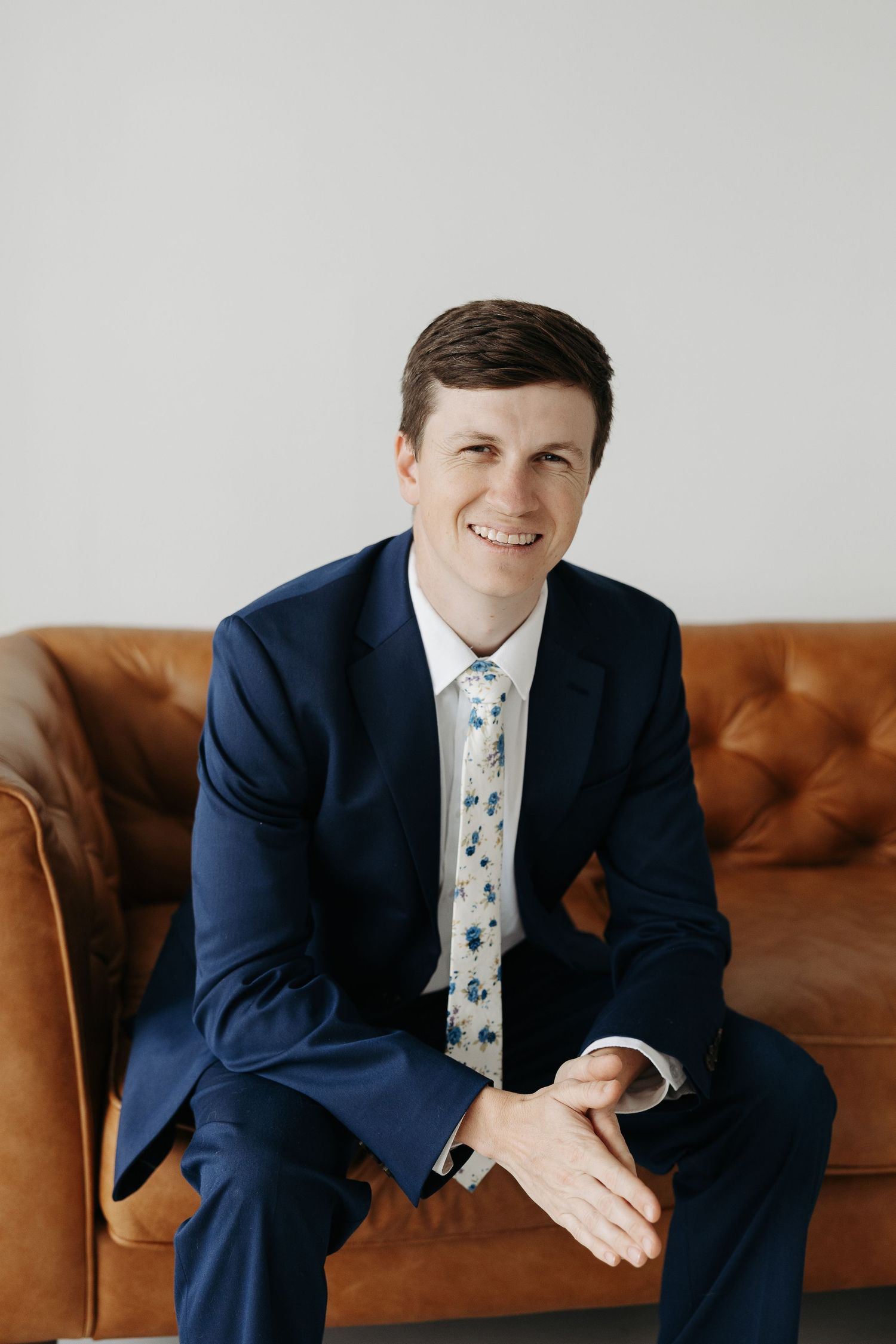 Professional headshot of person in navy suit sitting on brown leather couch.