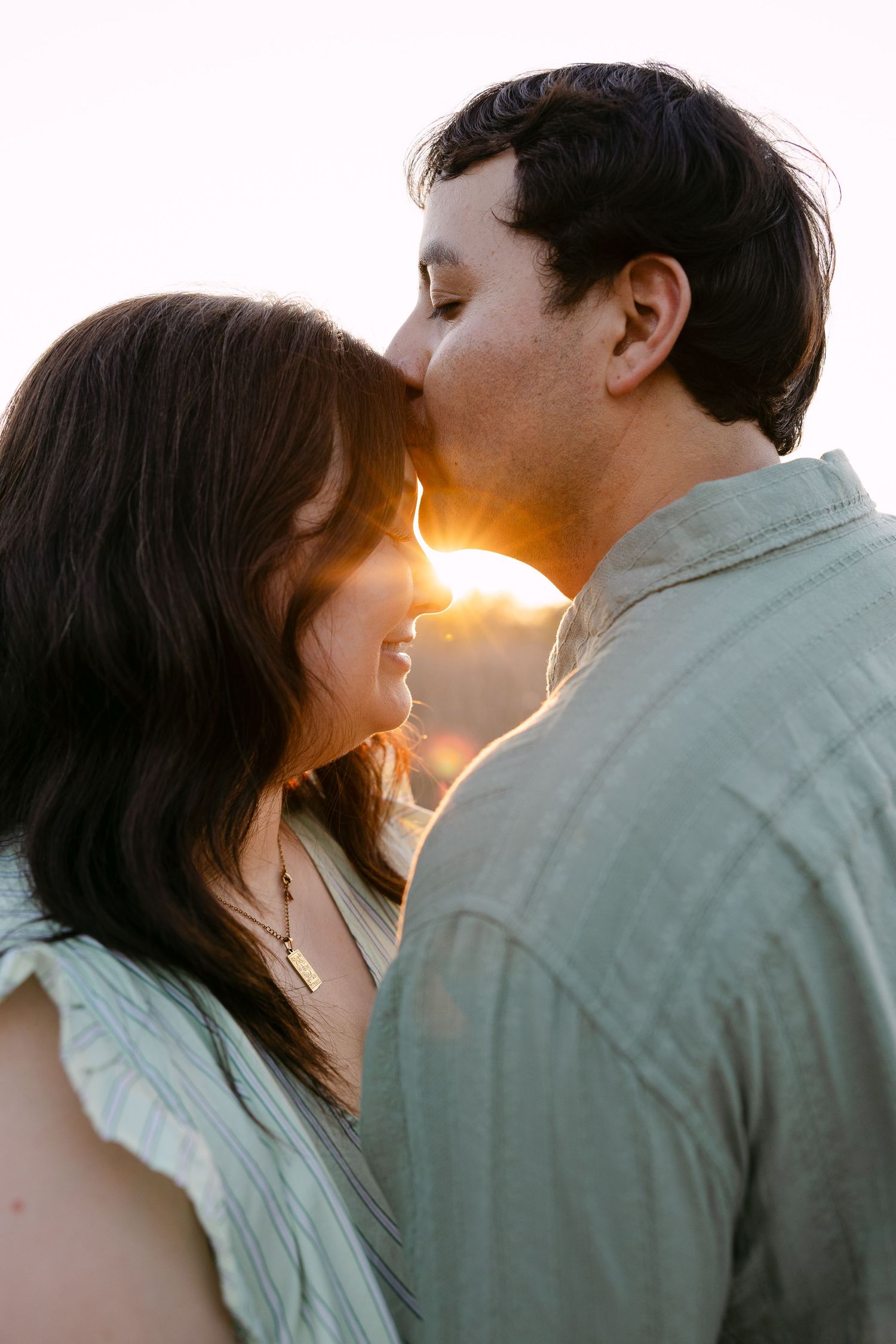 A couple shares a tender kiss at golden hour, backlit by warm sunset light in a romantic outdoor portrait.