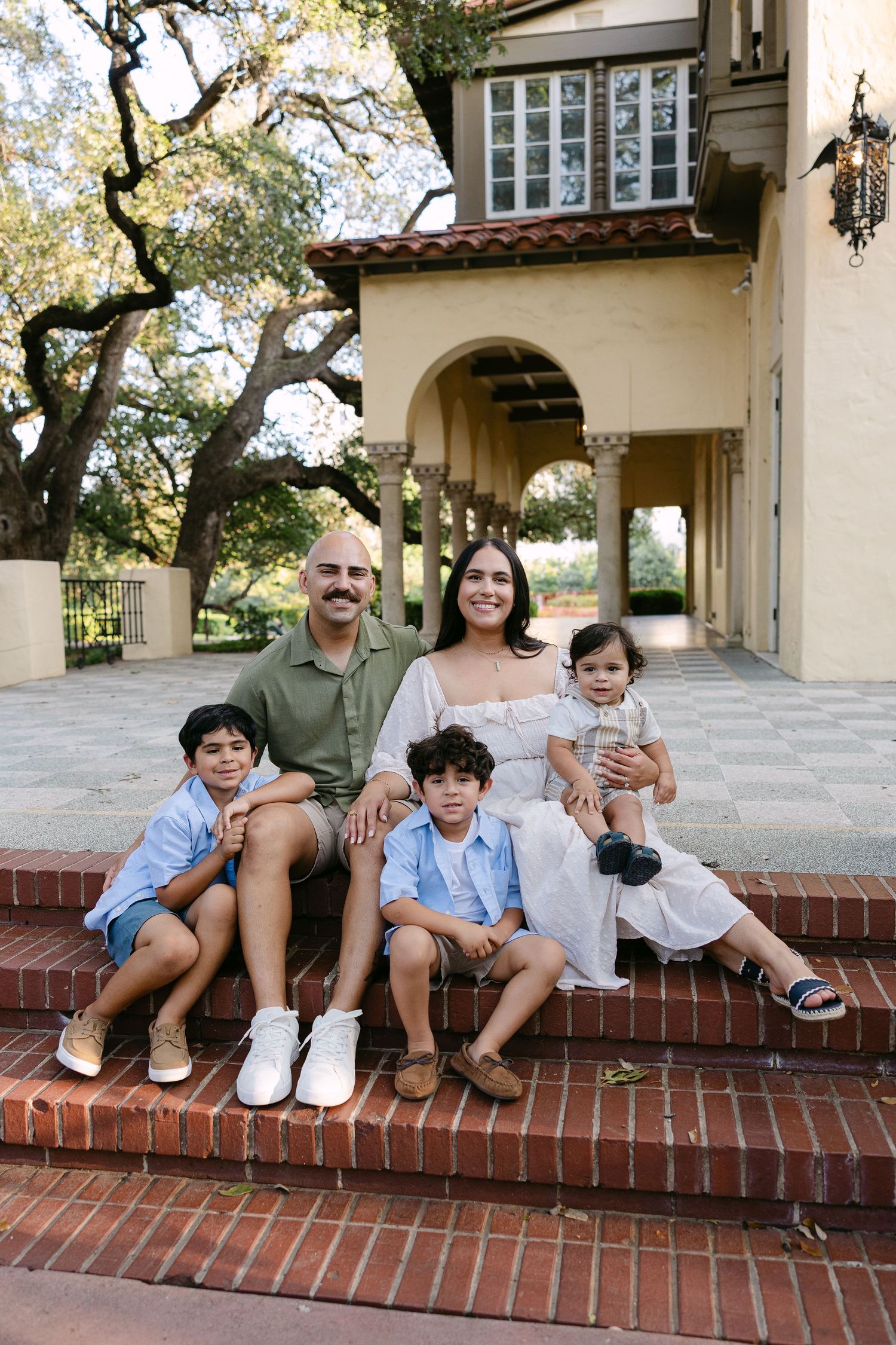 Family of five smiling on brick steps outside a Spanish-style home, parents with three young children in casual attire.