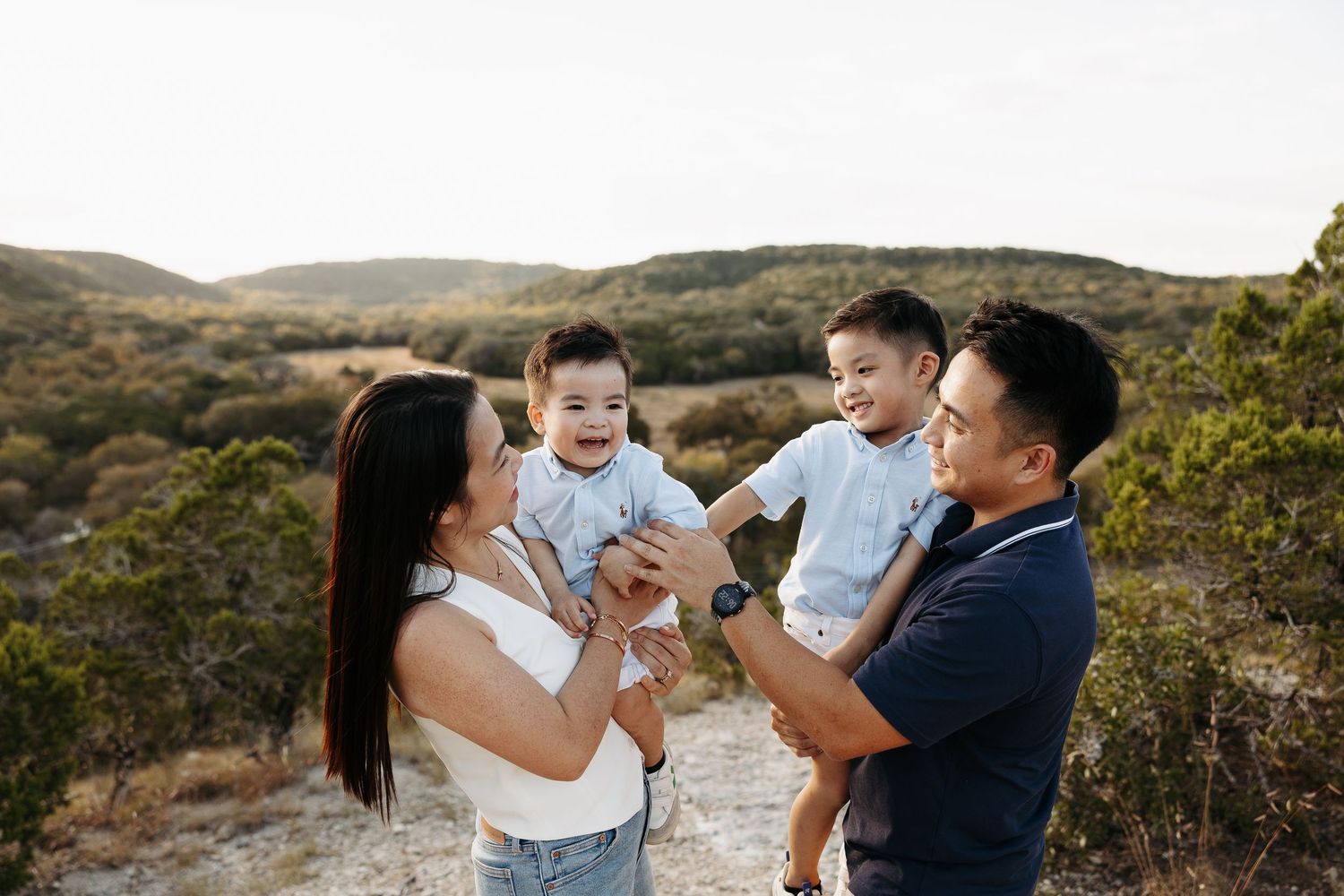 Asian family with two toddler boys smiling outdoors on rocky terrain with green hills in background.
