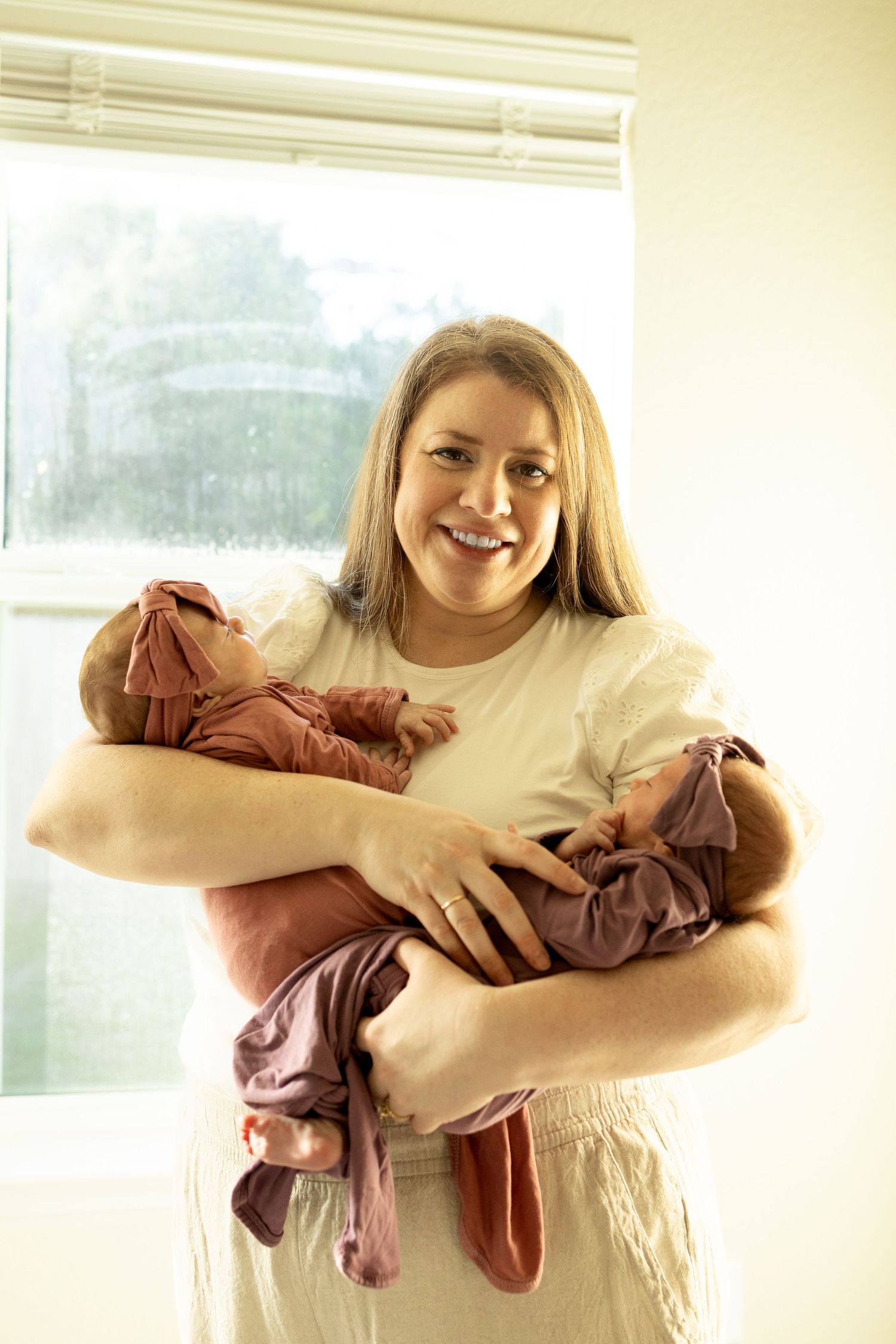 Woman in white dress holding infant wearing brown clothing in front of bright window.