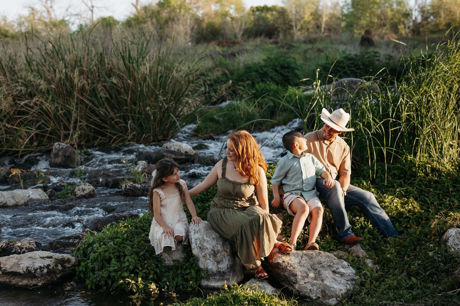 Family of four sitting on rocks by a creek in golden hour light, surrounded by lush greenery.