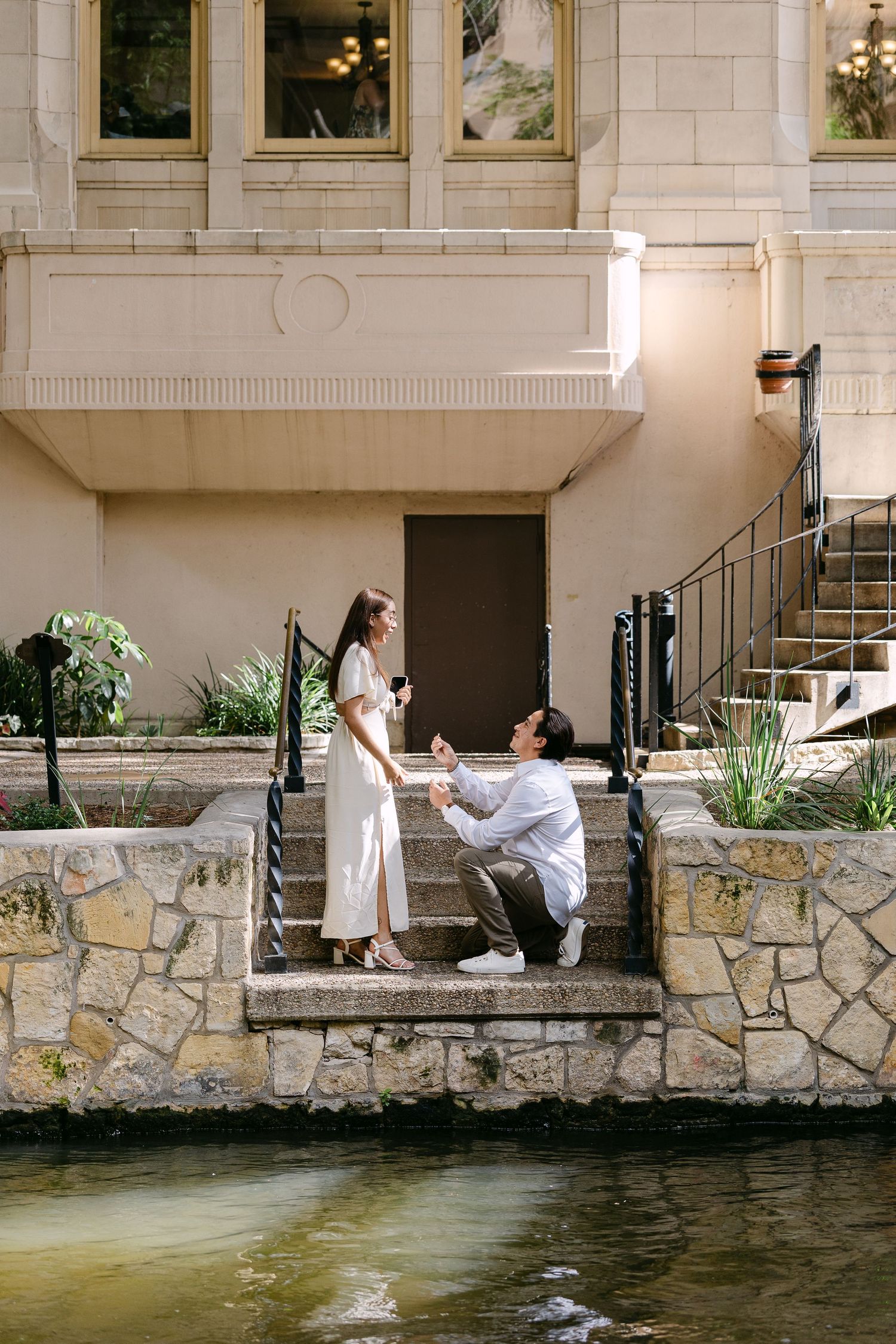Man proposes to woman on stone steps by a river near a elegant building, romantic outdoor marriage proposal scene.