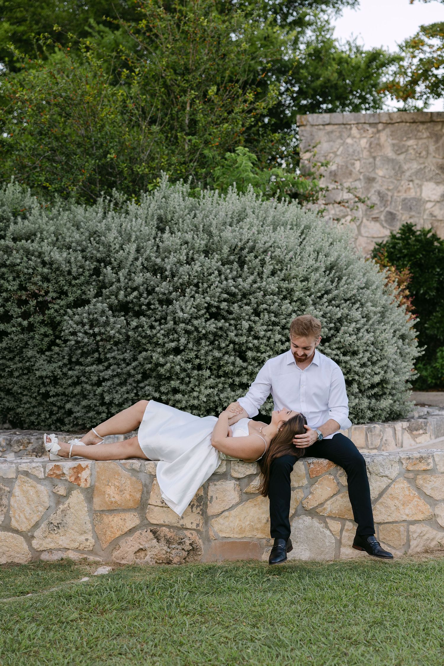 Couple posing on stone wall in garden, woman leaning back in white dress, man in white shirt and black pants.