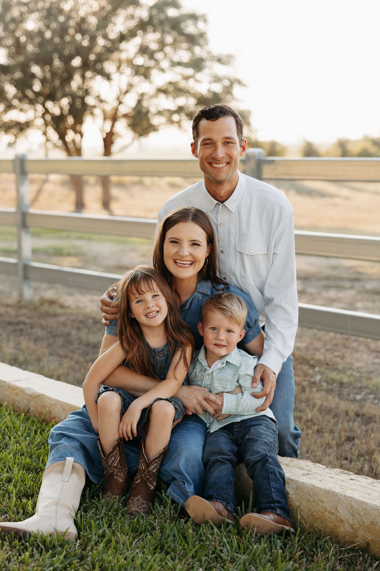 Family of four smiling outdoors near a wooden fence on a farm, parents with two young children sitting on grass.