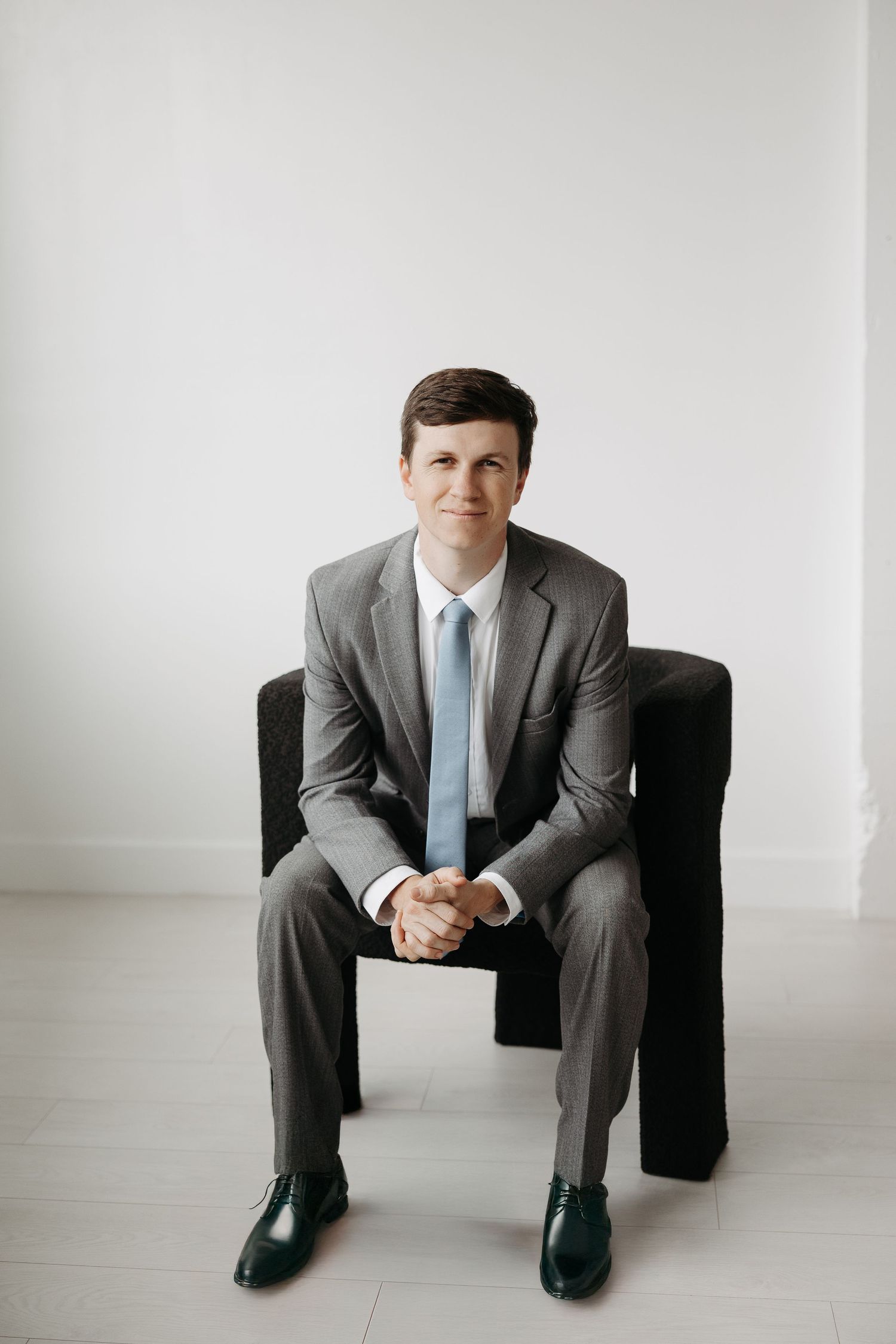 Professional portrait of person in grey suit seated on black chair against white background.