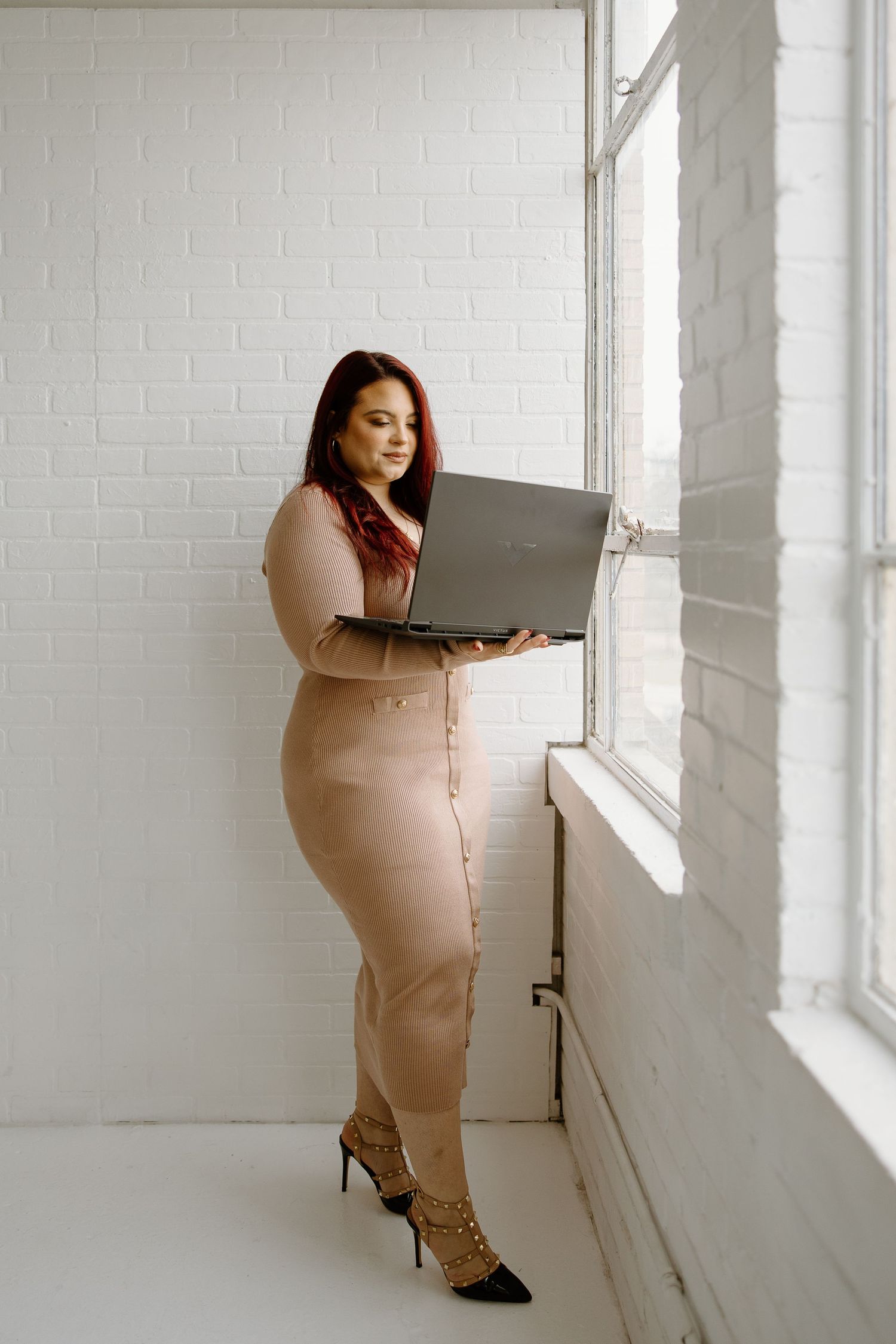 Professional wearing beige fitted dress works on laptop by bright window in modern office.