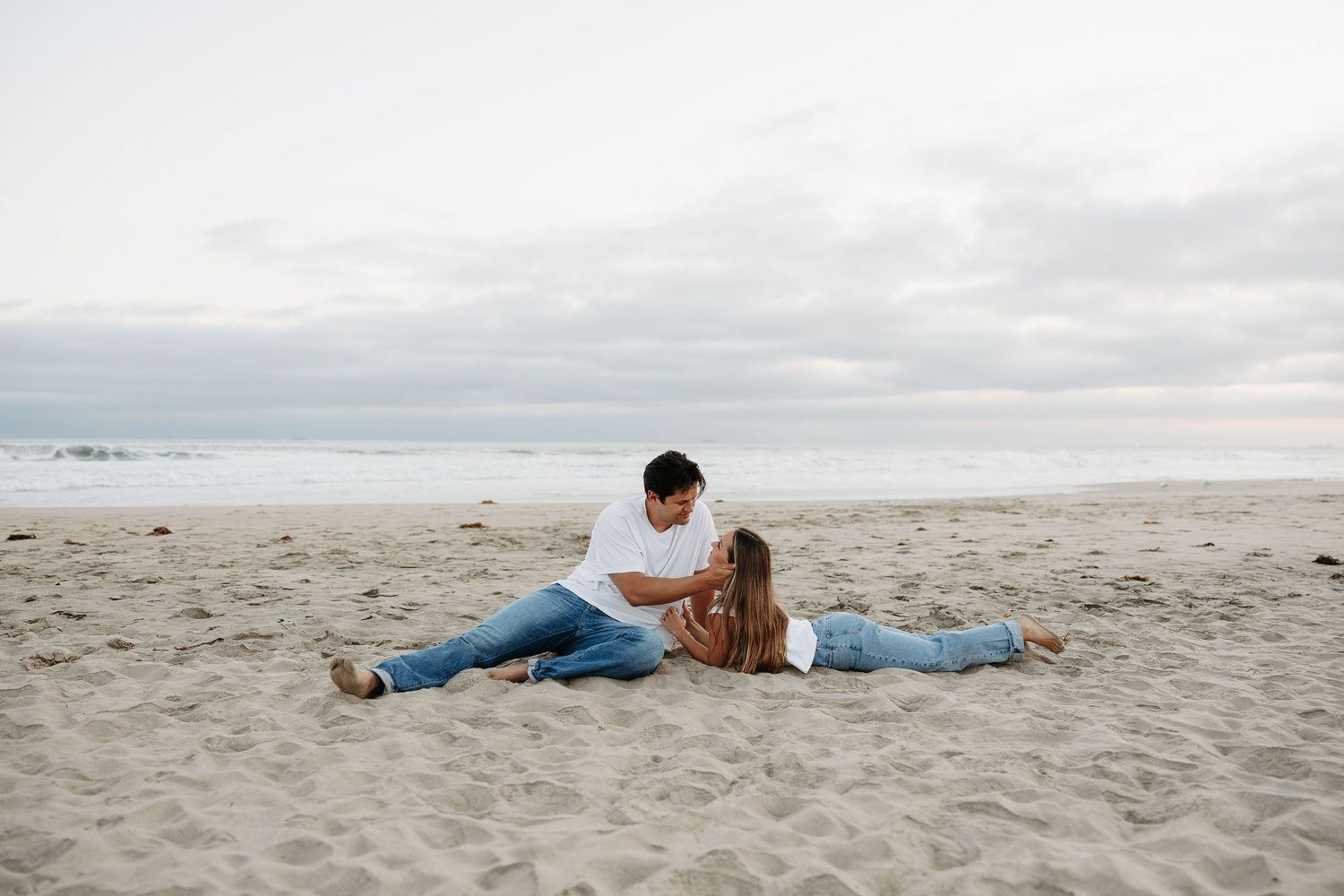 Couple sitting together on sandy beach, man looking down at woman lying across his lap, ocean waves in background.