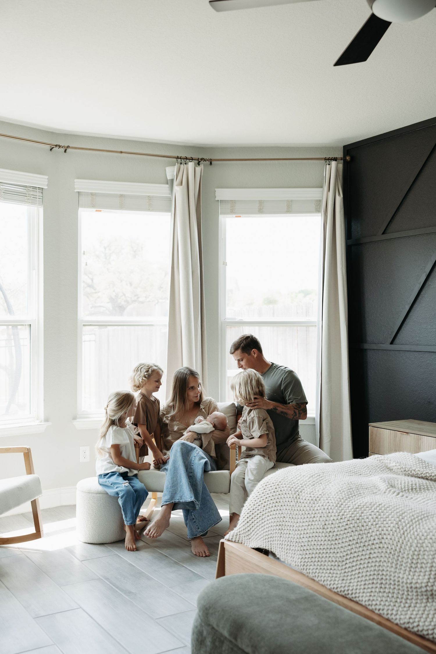 A family gathers in a bright bedroom, parents and children sharing a tender moment near bay windows with elegant curtains.