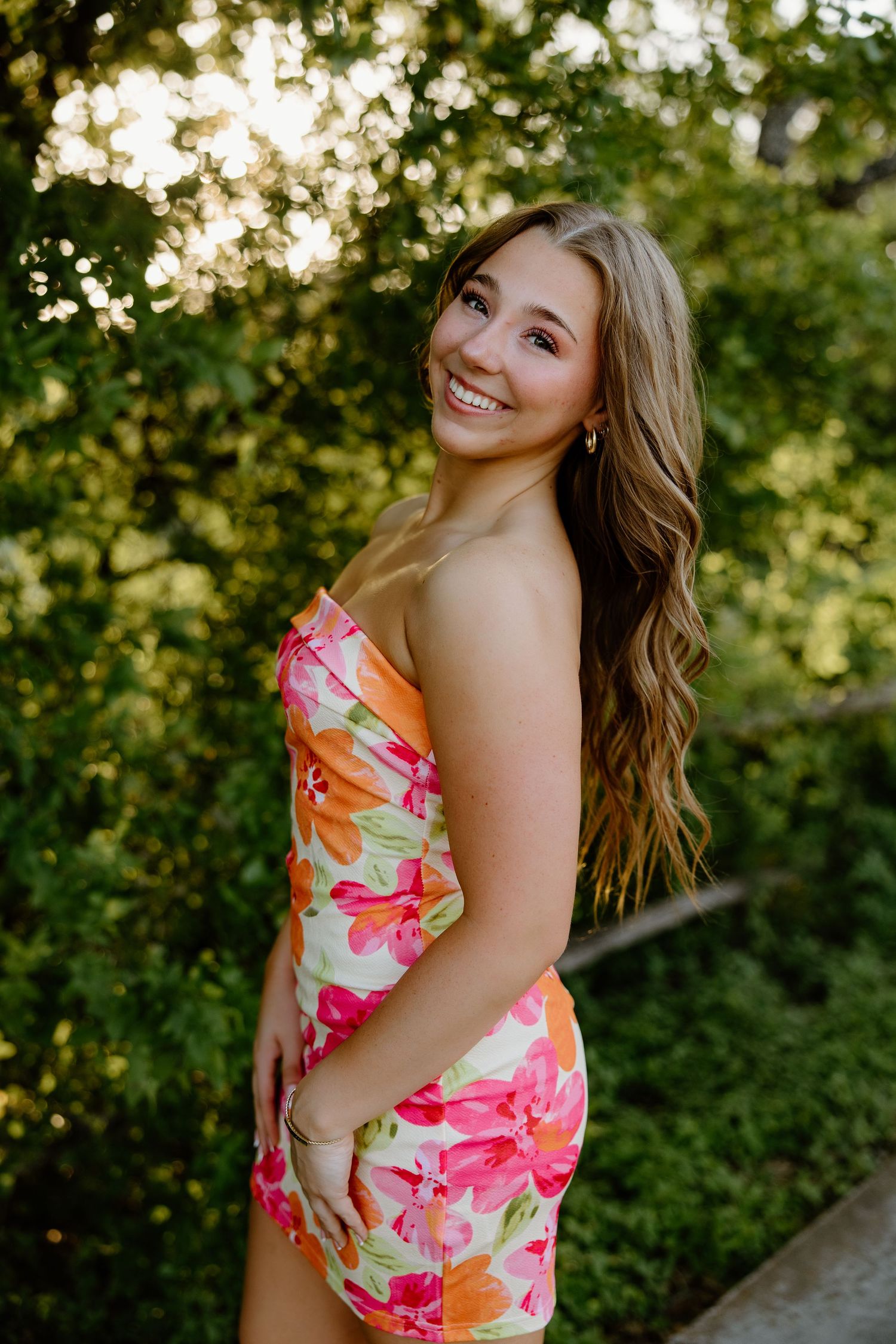 Woman in floral pink and orange dress smiles warmly during outdoor summer portrait session.