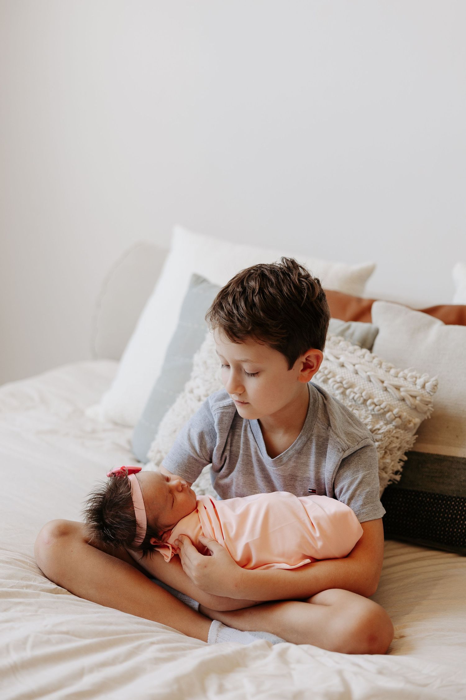 Two siblings share a tender moment on a white bed as the older brother gently holds and watches over the baby.