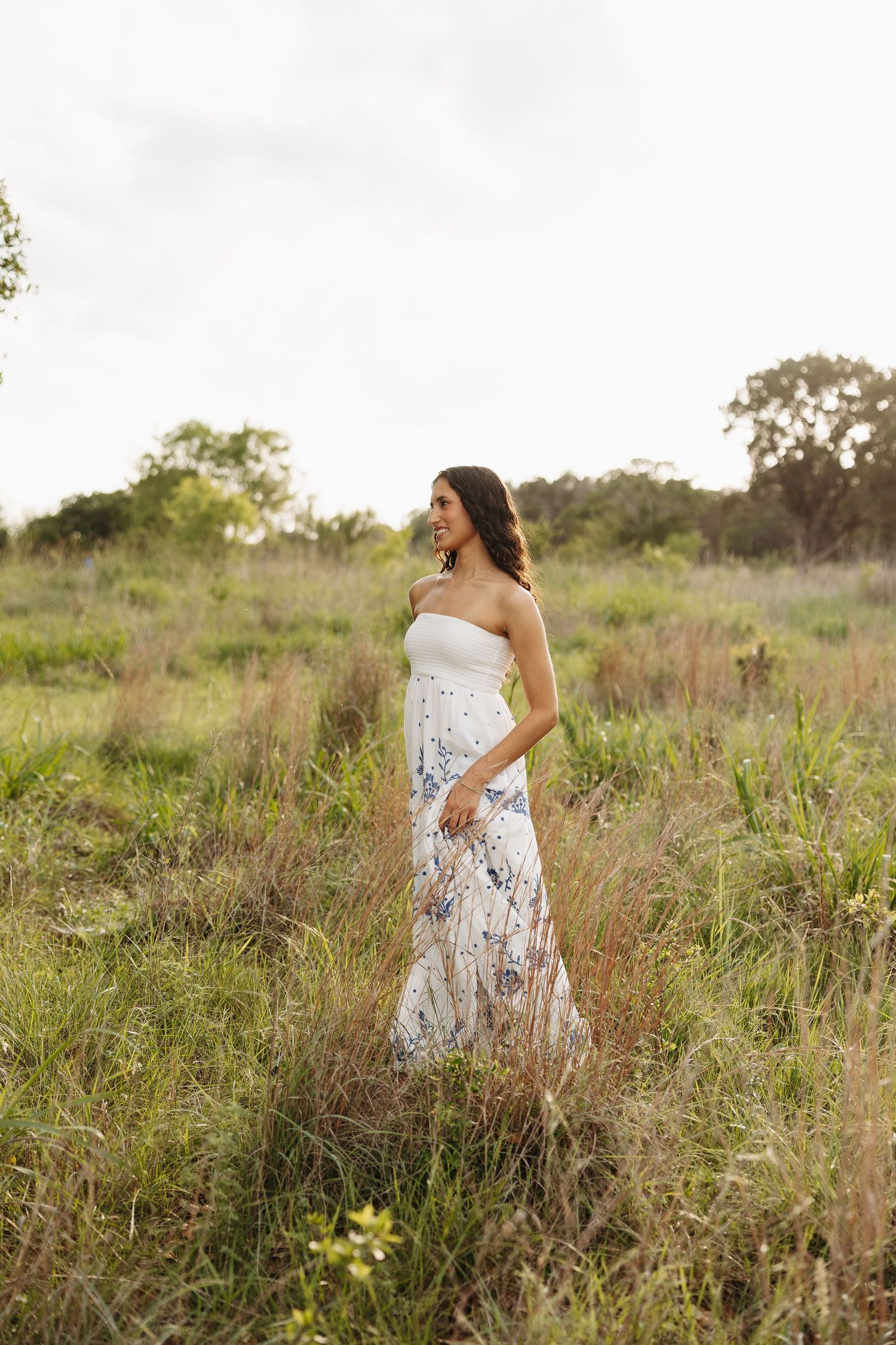 Woman in white lace dress poses gracefully in tall grass field during golden hour photography session.