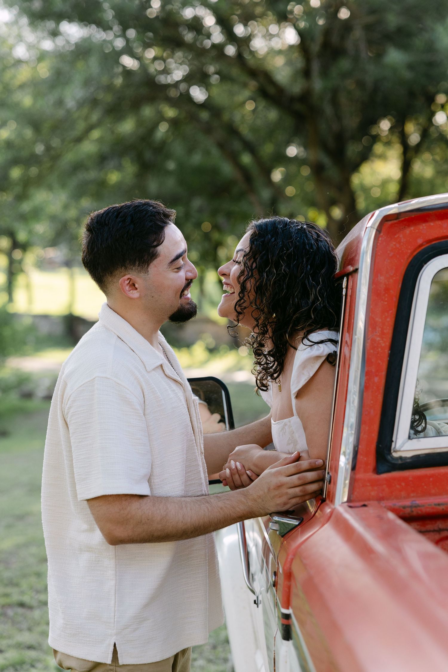 A couple shares a romantic moment beside a vintage red truck surrounded by lush green trees outdoors.