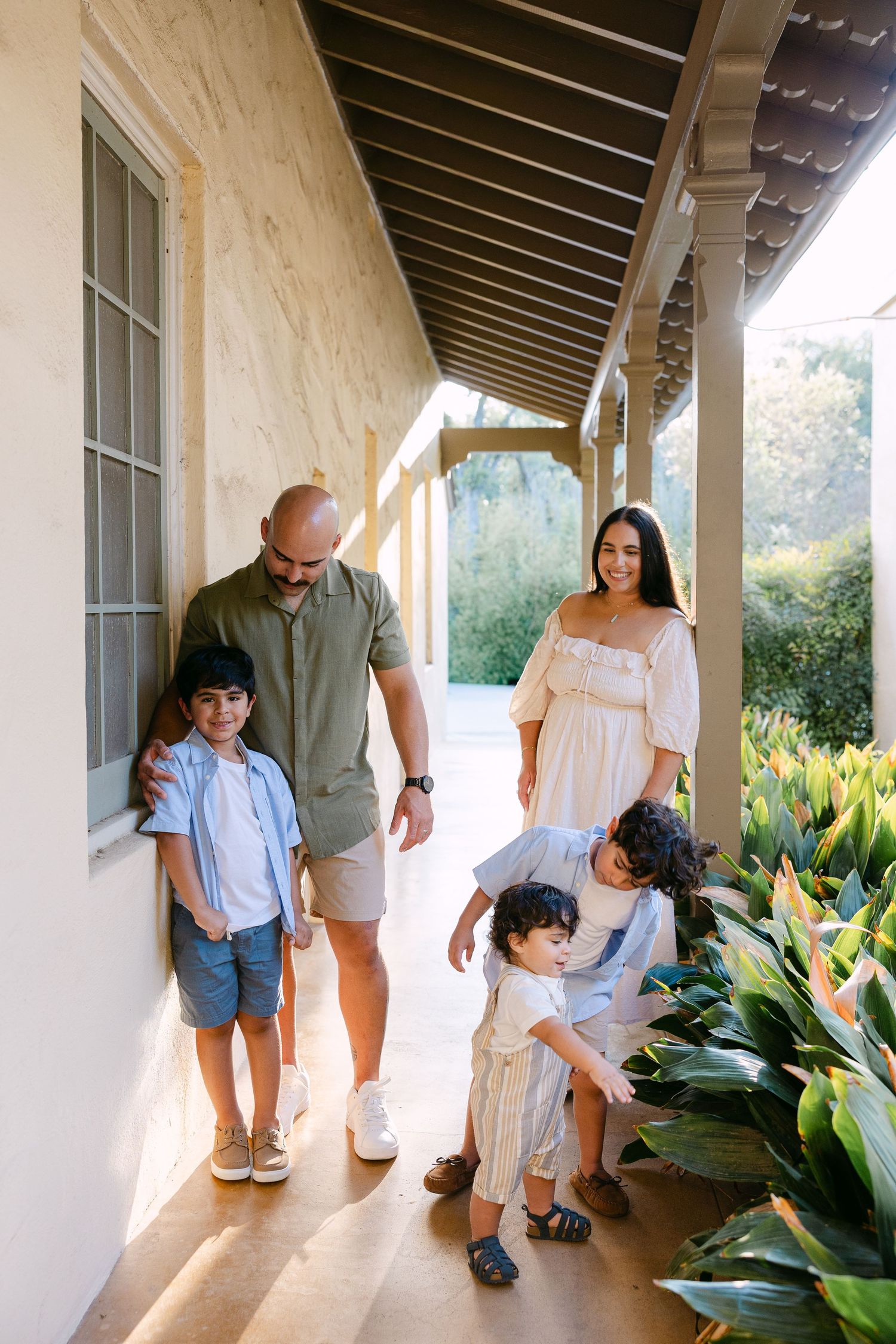 Family of five enjoying a sunny day on a covered porch, with parents and three children exploring lush tropical plants.