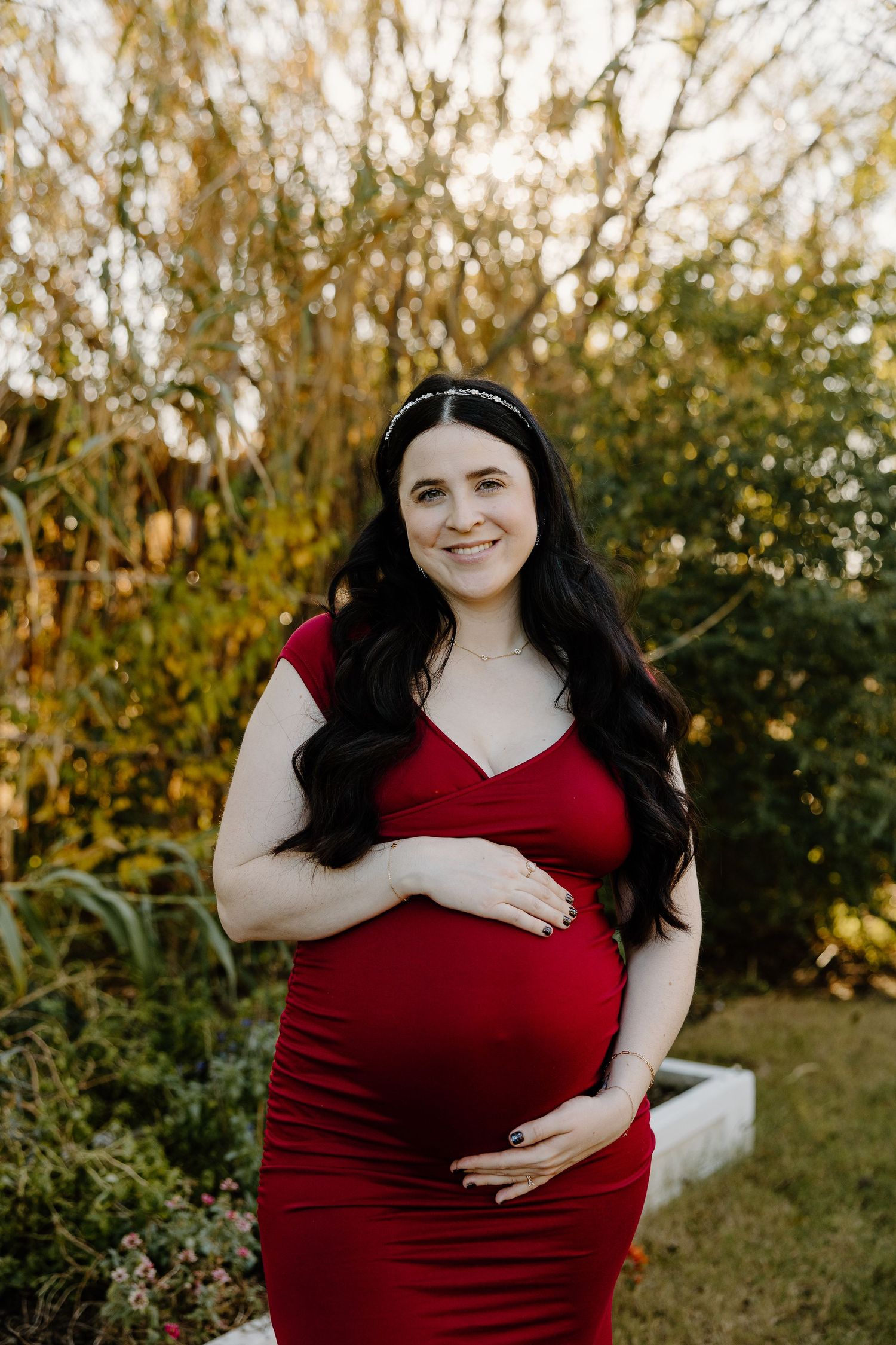 Expectant mother in red dress posing outdoors during sunset maternity photoshoot.