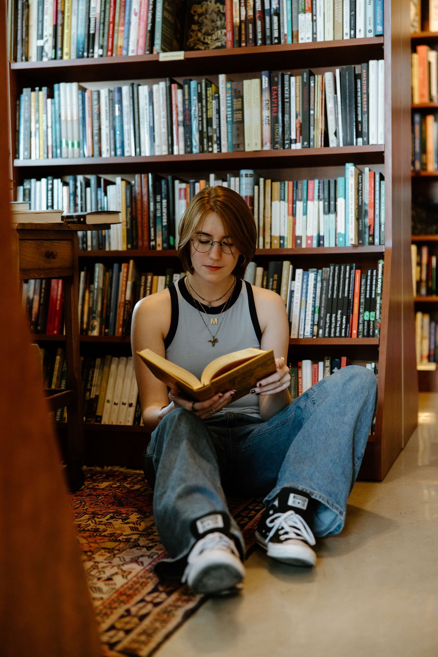 Student reading book while sitting on floor of library surrounded by bookshelves.