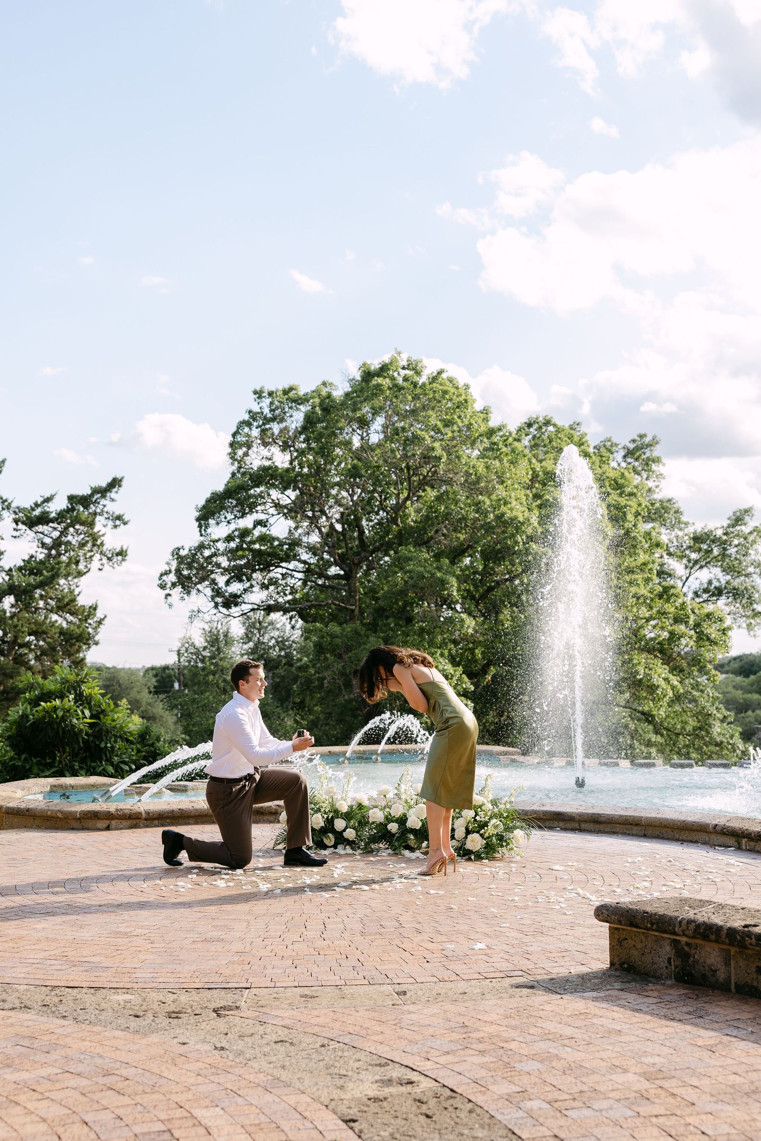 Man proposes on one knee by a fountain as woman leans forward emotionally, surrounded by flowers in a garden.