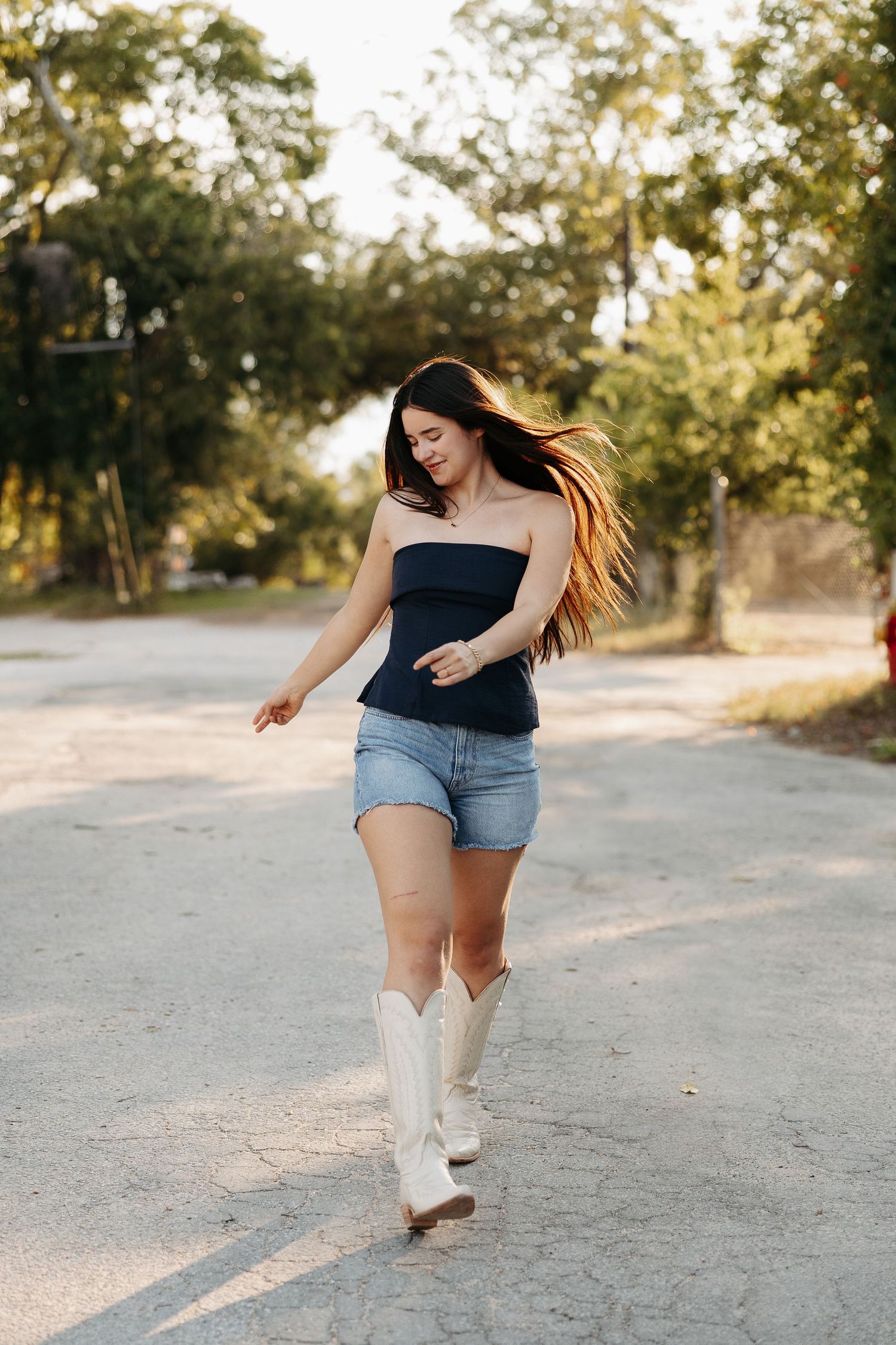 A woman in a black top and denim shorts dances on a dirt path wearing white cowboy boots at sunset.