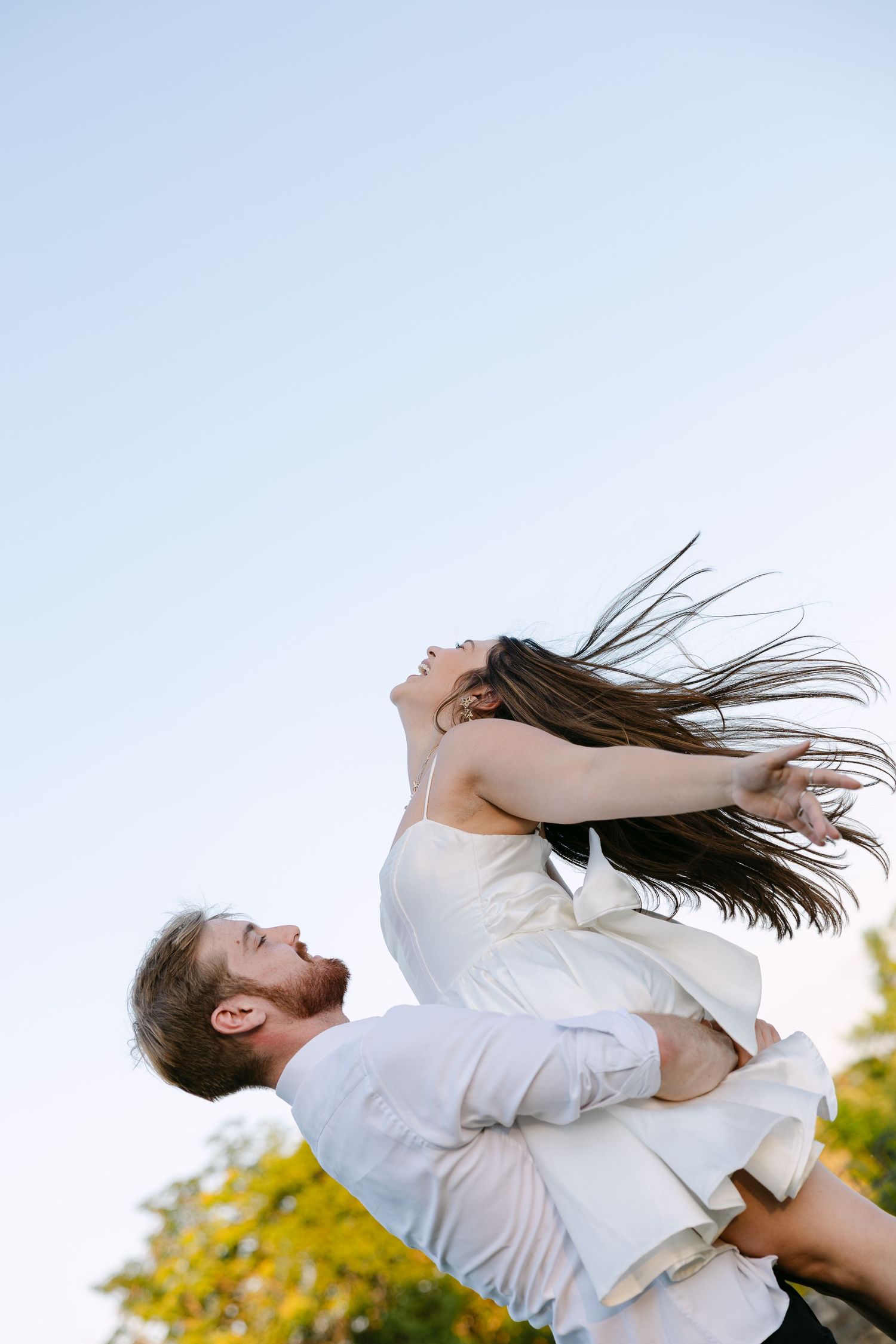 A joyful groom lifts his bride outdoors, her long hair flowing in the wind against a clear blue sky.