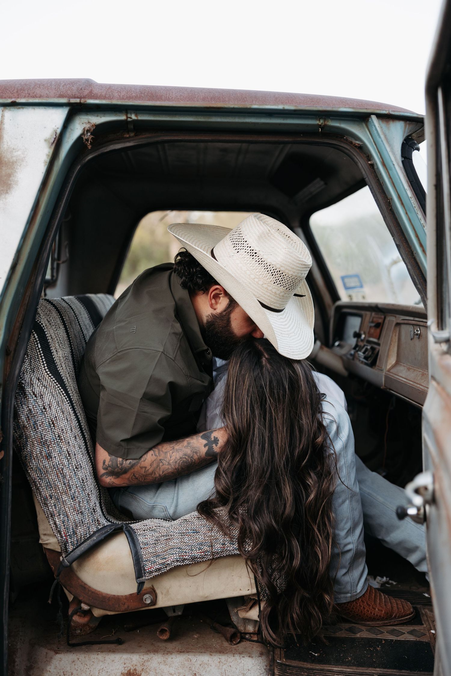 A bearded man in a white cowboy hat kisses a woman with long dark hair inside a vintage truck cab.