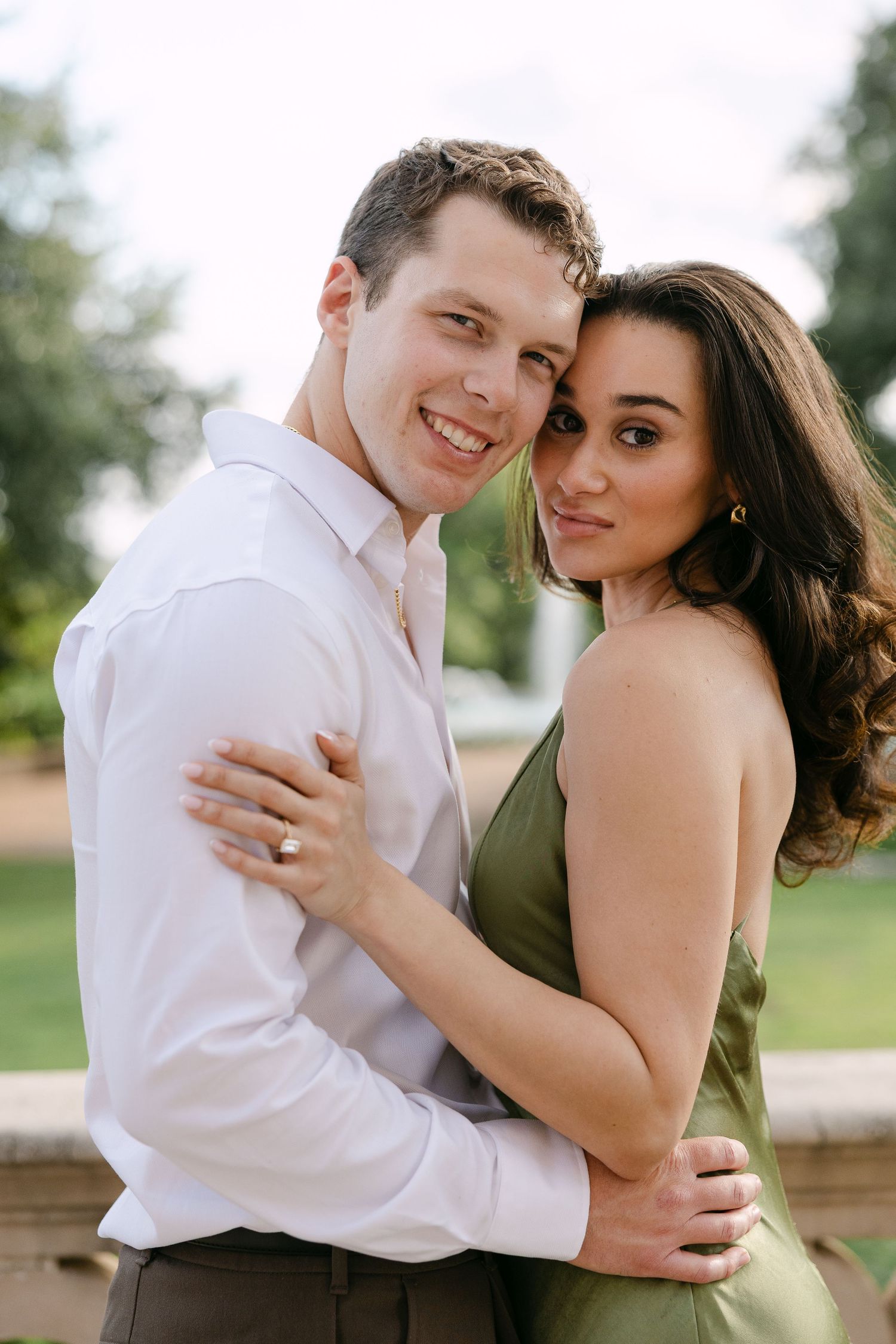 A smiling couple embraces outdoors, the woman in a green dress and the man in a white shirt, posing for an engagement photo.