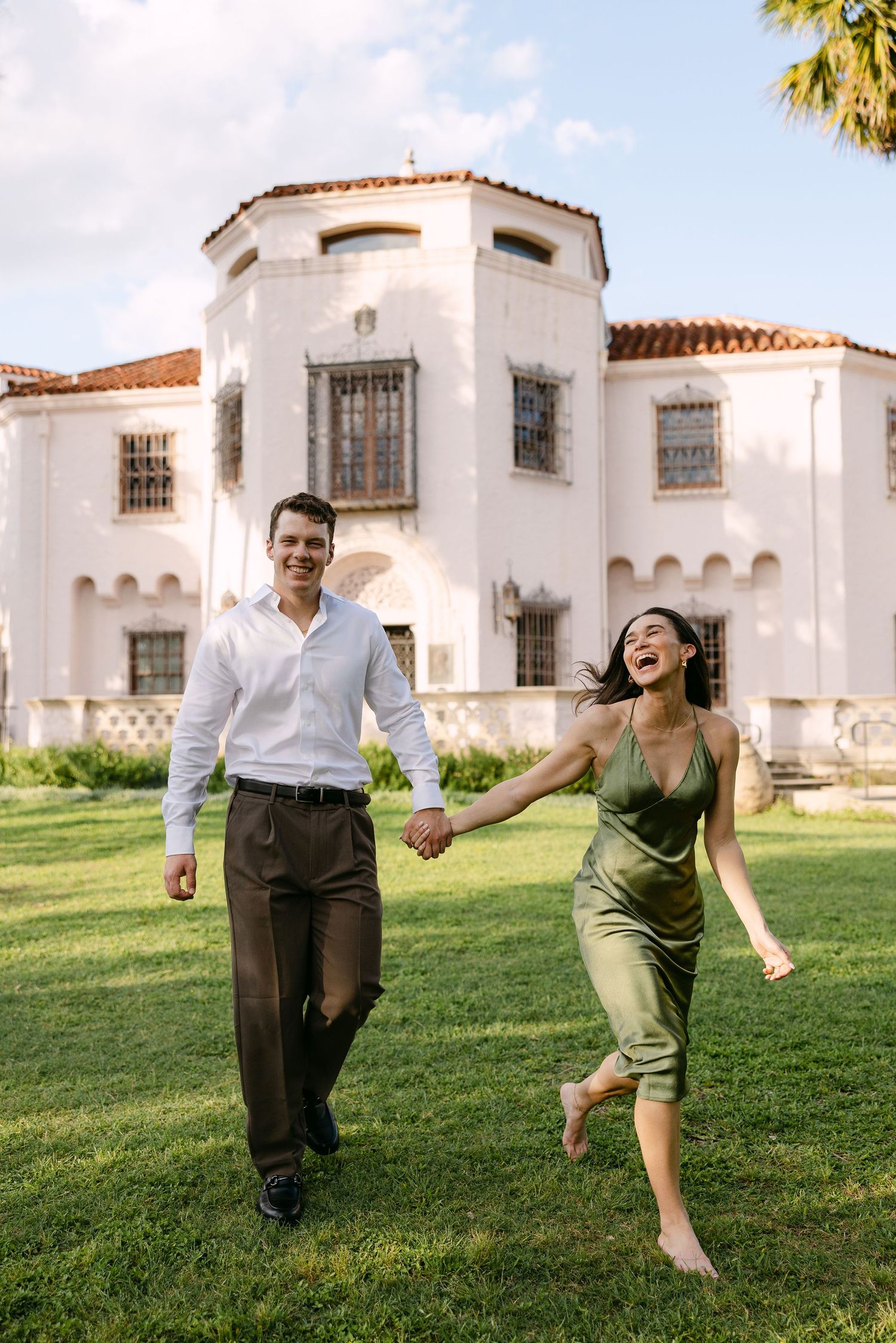 A joyful couple holds hands and runs across a green lawn in front of a white Spanish-style villa.