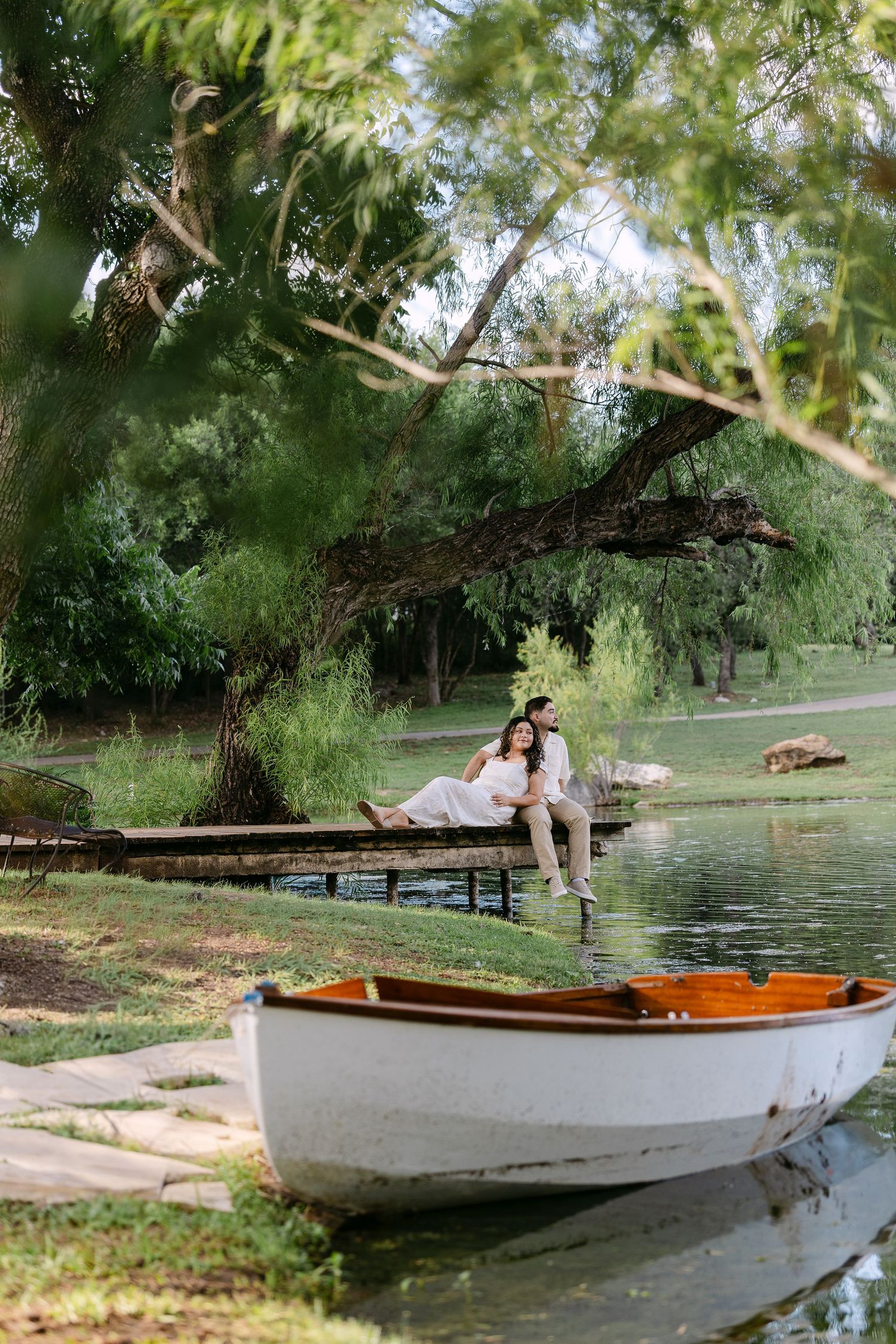 Couple sitting on a wooden dock by a serene lake, surrounded by lush green trees, with a white boat in the foreground.
